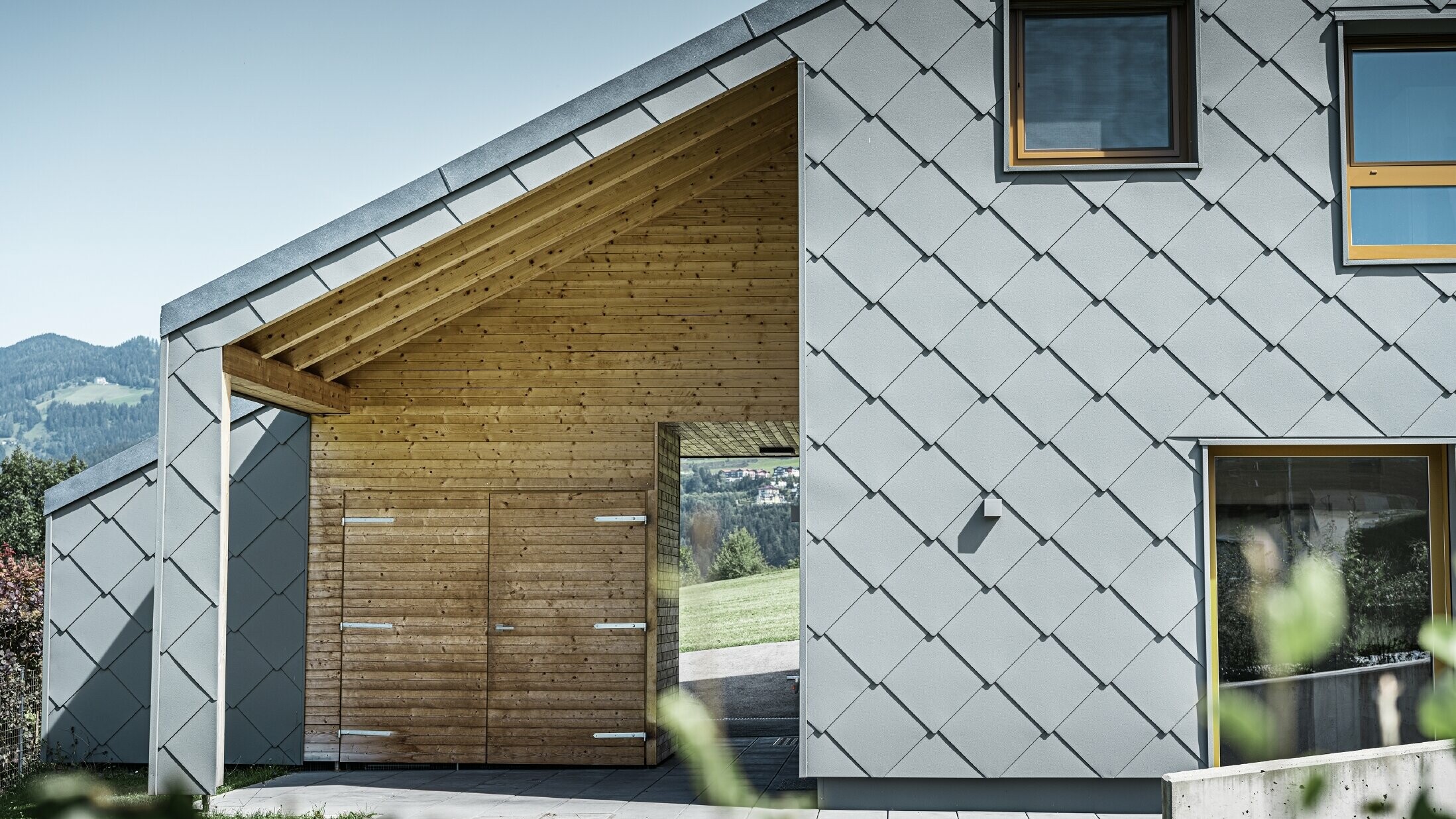 L'ingresso coperto è realizzato con un rivestimento orizzontale in legno, mentre il resto della facciata è rivestito con la scaglia per facciata PREFA grande in alluminio grigio chiaro.