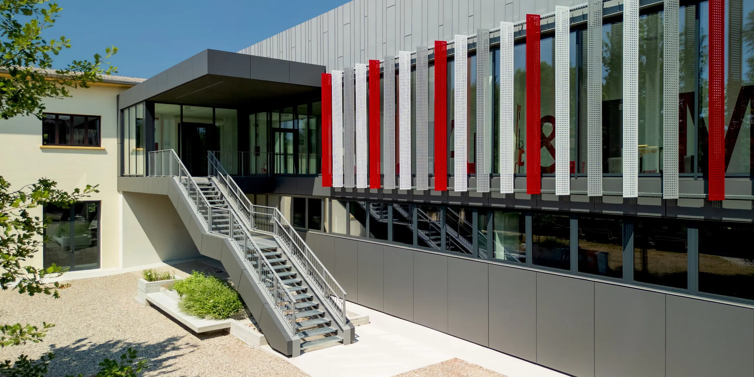 La cour intérieure accueillante de l'école d'art et de métiers F. Bertazzoni à Suzzara est agrémentée d'un élégant escalier extérieur menant à l'étage supérieur vitré. La façade en aluminium PREFA gris sombre, sur deux niveaux, séduit par le jeu dynamique d'éléments perforés gris clair et de bandes rouge vif, derrière lesquelles on aperçoit une inscription grand format. Les produits PREFALZ et PREFABOND de PREFA impressionnent par leur fabrication précise, leur qualité de surface durable et leur excellente résistance aux intempéries, conférant à l'établissement une identité forte et un design affirmé.