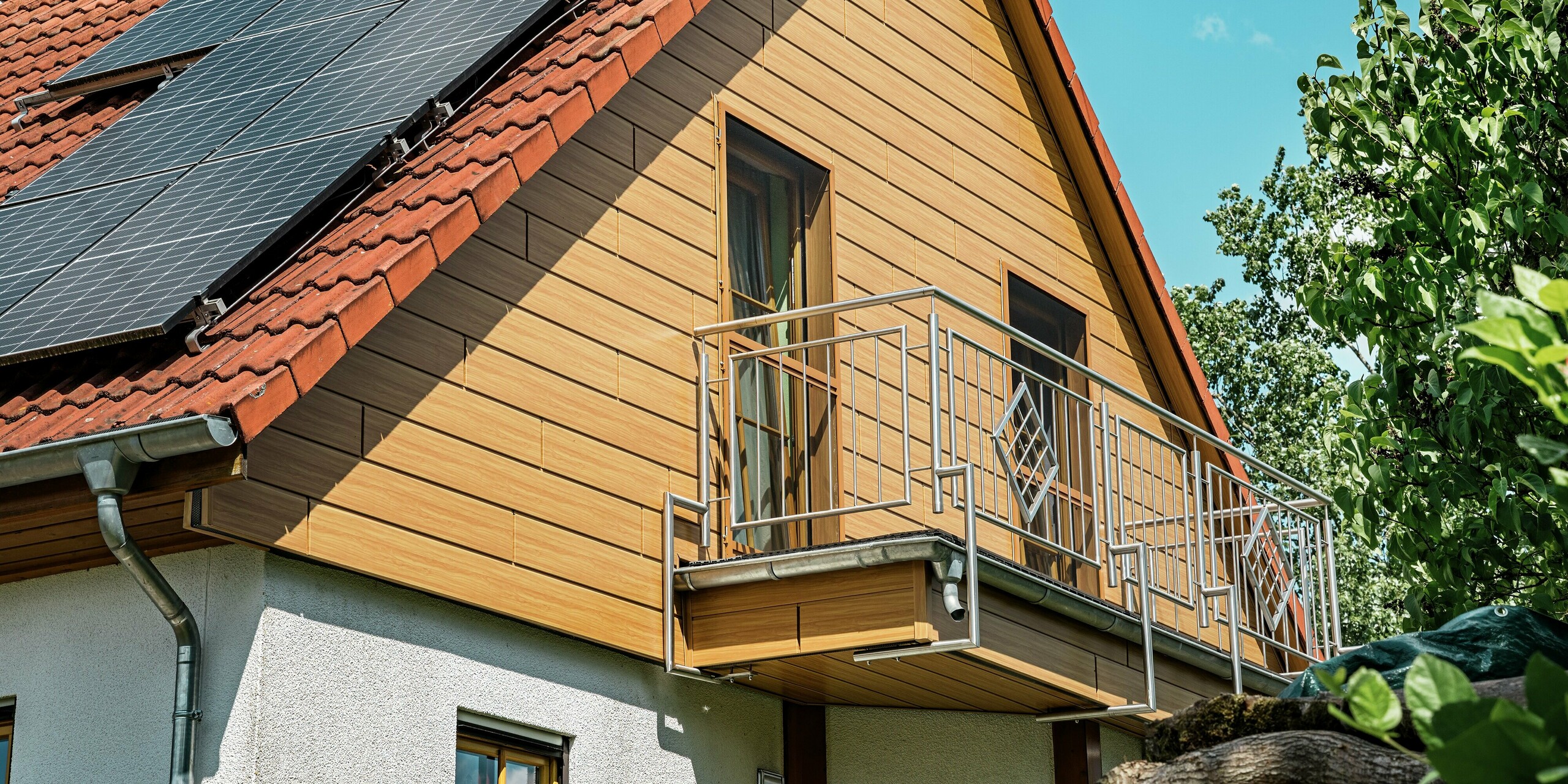 L'image présente le pignon récemment rénové d'une maison individuelle à Blieskastel, revêtu de Siding PREFA en chêne naturel. L'aspect chaleureux du bois, obtenu grâce à la robustesse de l'aluminium, s'harmonise parfaitement avec la toiture en tuiles rouges et l'imposant système photovoltaïque qui s'étend majestueusement sur toute la surface. Le délicat balcon métallique souligne l'esthétique moderne et crée un contraste saisissant avec la texture naturelle de la façade. Grâce à la surface PREFA résistante aux intempéries et inaltérable, ce revêtement conservera son aspect impeccable pendant de nombreuses années, un véritable atout pour l'ensemble du bâtiment.