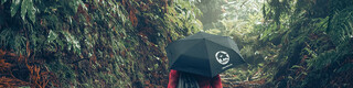 Photo : promeneuse en forêt, en veste rouge avec parapluie et sac à dos PREFA, symbolisant la protection de l’environnement et la durabilité PREFA, ainsi que l’économie circulaire et le recyclage