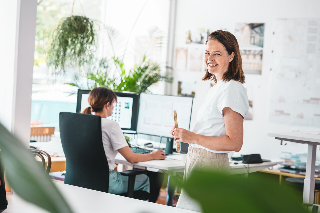Zwei Architektinnen im modernen Büro: Eine arbeitet am Computer mit digitalen Plänen, während die andere mit Maßstab und Entwürfen bereitsteht. Das Bild steht für effizienten Zugriff auf Planungsunterlagen und technische Ressourcen im PREFA Downloadcenter für Architekten und Planer.