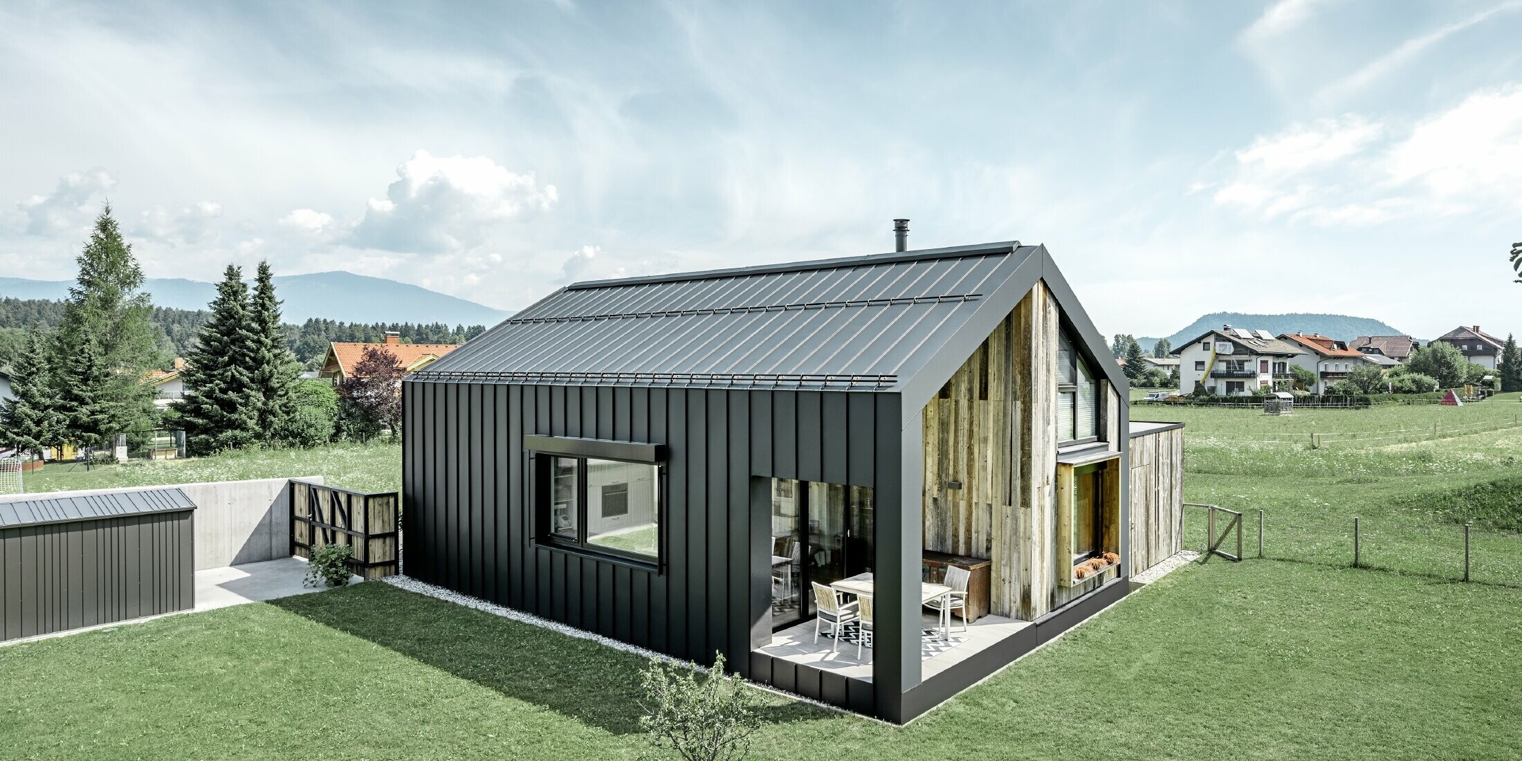 Maison individuelle avec toit à pignon ; la toiture et la façade sont recouvertes avec PREFALZ de PREFA en P.10 anthracite. Les bacs de la surface de toit se prolongent jusque sur la façade. Une partie de la façade est revêtue de bois patiné.