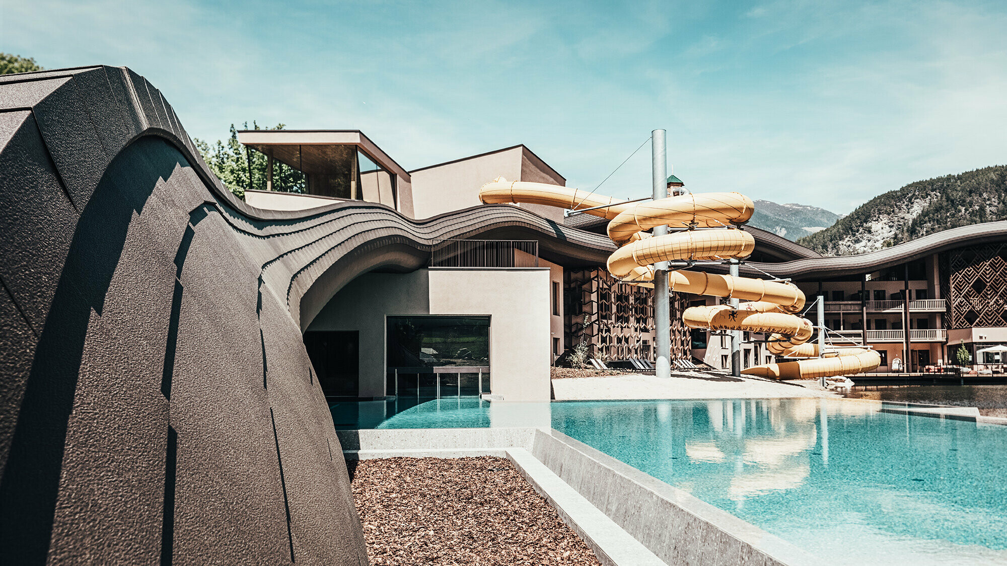 Aperçu de la piscine de l'hôtel Falkensteiner, en arrière-plan on voit le toboggans jaunes. La vague en bardeaux de façade prefa en P.10 brun dominent le complexe.