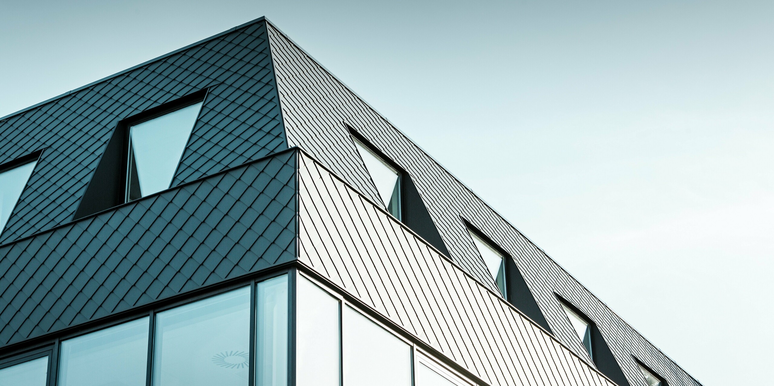 Das moderne Bürogebäude Kasteeltje Heizel in Strombeek-Bever nahe Brüssel verfügt über eine markante PREFA Aluminium-Fassade aus anthrazitfarbenen Wandrauten. Die Nahaufnahme der geometrischen Rautenstruktur bildet einen spannenden Kontrast zu den großen Glasflächen und verleiht dem Gebäude eine dynamische, futuristische Ausstrahlung. Die langlebigen und wartungsarmen PREFA Aluminiumprodukte vereinen Ästhetik, Nachhaltigkeit und Funktionalität in einem architektonischen Eyecatcher.