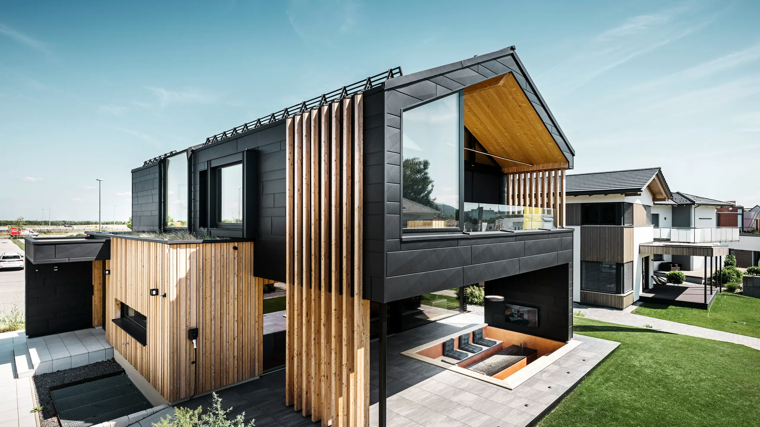 Vue latérale de la maison modèle Autark dans le Blue Lagoon à Vösendorf, en Autriche. L'étage supérieur impressionne par ses grandes surfaces vitrées et sa façade moderne en aluminium composée de panneaux PREFA Siding.X en P.10 noir, associés à des éléments verticaux en bois. La terrasse spacieuse au rez-de-chaussée avec sièges encastrés s'intègre parfaitement dans l'architecture. Le design élégant et durable de la maison combine de manière impressionnante l'aluminium et le bois.