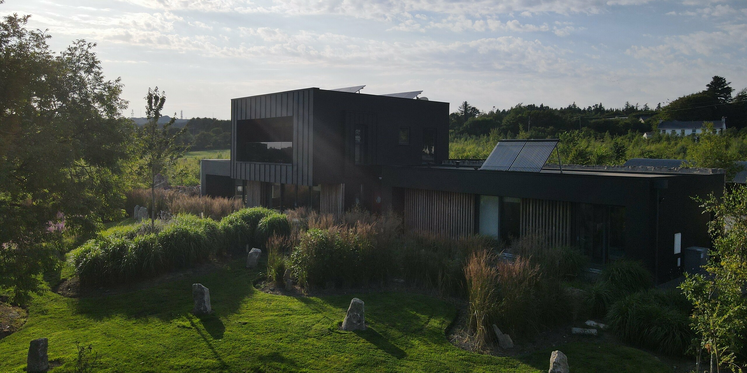 La photographie présente une maison individuelle moderne à Wexford, nichée au cœur d'un jardin luxuriant et paysagé. La façade noire PREFALZ, coloris P.10 noir, est particulièrement saisissante : ses lignes épurées et son élégant aspect métallique contrastent avec la verdure éclatante. La lumière intense du soleil couchant accentue la texture du revêtement en aluminium et souligne ses propriétés de haute qualité et sa résistance aux intempéries. L'alliance architecturale de formes cubiques modernes, de grandes fenêtres et du matériau PREFA robuste fait de ce bâtiment un véritable emblème architectural.