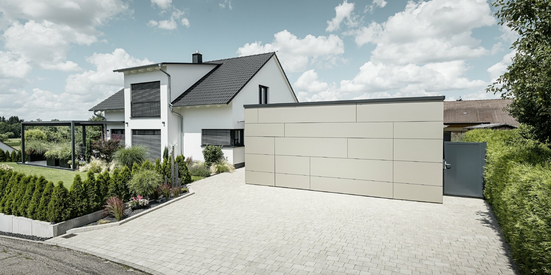 La façade du garage de cette maison individuelle est entièrement revêtue avec le panneau composite en aluminium couleur bronze. On ne distingue pas non plus la porte de garage des joints. Une allée spacieuse s'étend juste devant.