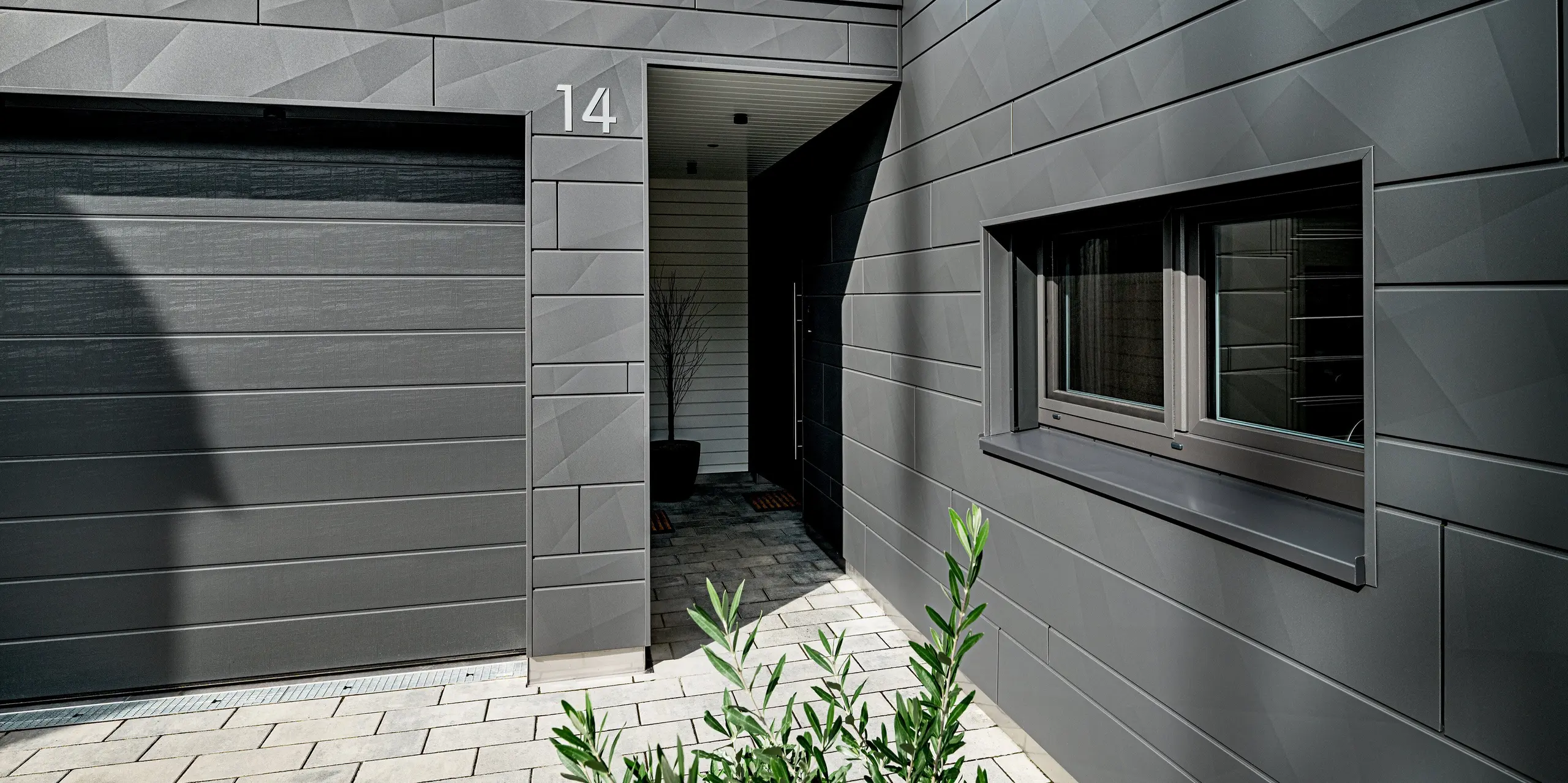 Entrée et garage d'une maison individuelle moderne à Heidenheim, revêtue d'une façade en aluminium Siding.X PREFA couleur P.10 gris sombre. Les bordures travaillées des panneaux créent un motif géométrique dynamique. Le numéro « 14 » est clairement visible sur l'élément de façade au-dessus de l'entrée couverte. À droite, une grande fenêtre aux contours précis domine la scène, tandis que l'allée pavée et l'aménagement paysager discret complètent l'ensemble résolument moderne. La surface PREFA, résistante et durable, offre une protection optimale et une stabilité à toute épreuve.