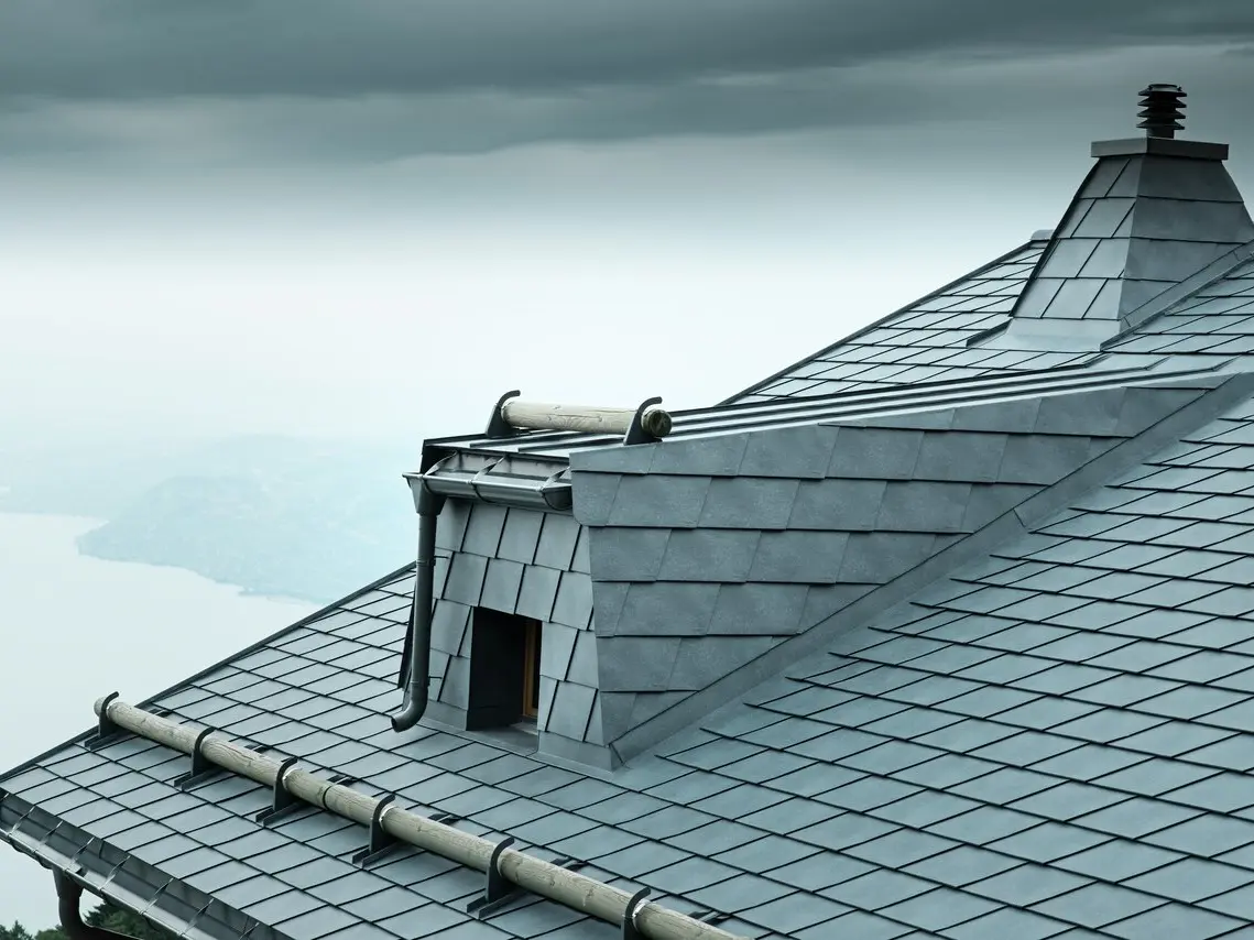 Vue du toit d'un chalet en bois au bord du lac Léman avec bardeaux PREFA gris pierre, lucarne, pare-neige et cheminée. La photo montre une construction de toit moderne et fonctionnelle pour une architecture durable dans les régions alpines.
