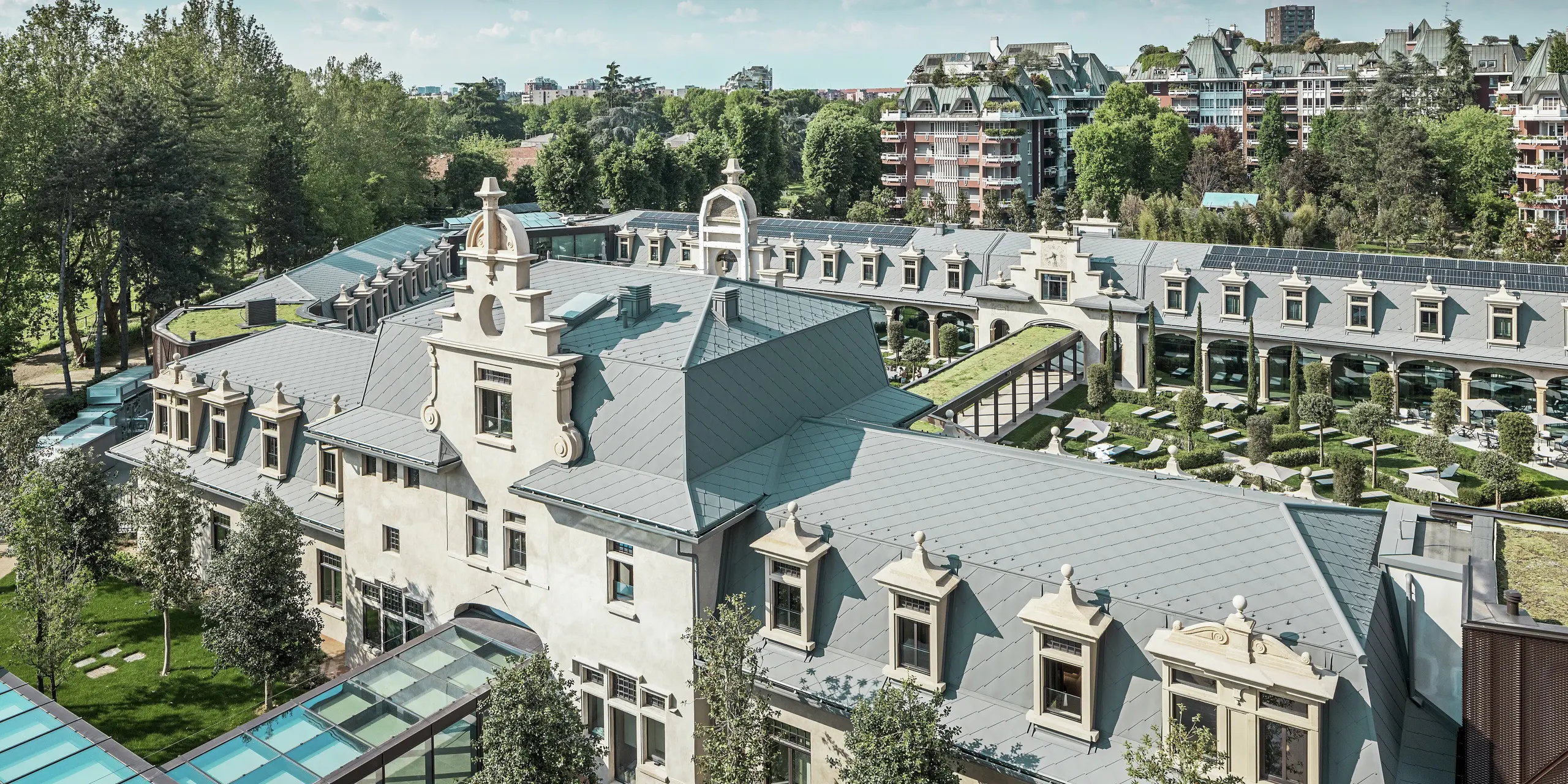 Vista panoramica dell'imponente complesso “De Montel – Terme Milano” con architettura Art Nouveau protetta come monumento storico e moderno ambiente benessere. L'ampia copertura del tetto con scaglie PREFA 44 × 44 in P.10 grigio chiaro conferisce al complesso un'elegante uniformità. I moduli fotovoltaici integrati, i tetti verdi e i collegamenti in vetro tra gli edifici riflettono l'esigenza di sostenibilità e design contemporaneo nel contesto urbano.