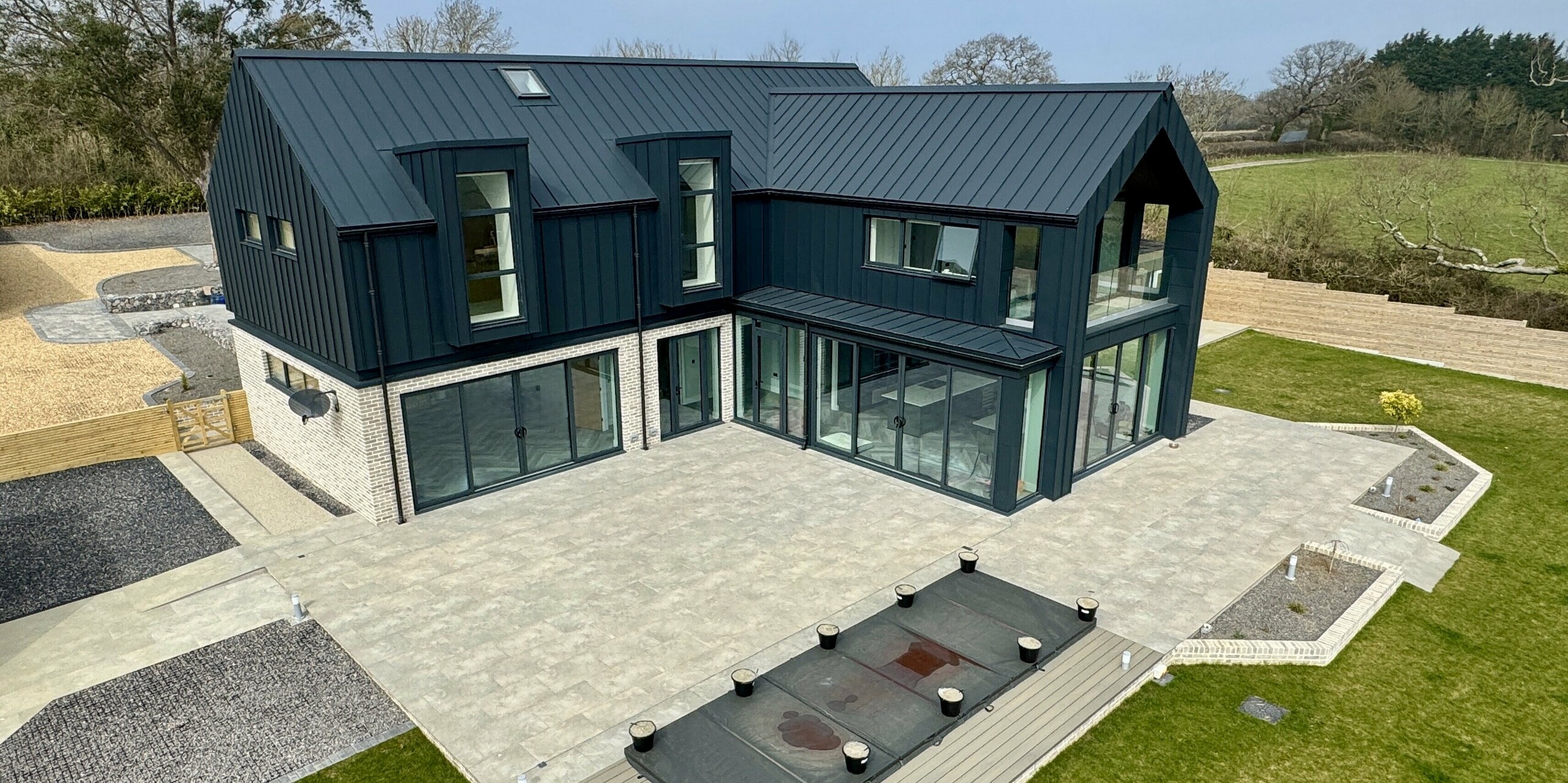 Maison individuelle moderne avec une toiture et une façade PREFALZ remarquables en P.10 gris sombre, associées à un parement en pierre claire au rez-de-chaussée. De larges baies vitrées ouvrent l'espace de vie sur une vaste terrasse avec piscine. Les surfaces en aluminium sont durables, résistantes à la corrosion et parfaitement adaptées au climat côtier. Le jeu de contrastes entre la toiture foncée, le verre et la pierre naturelle crée une élégante harmonie et souligne la qualité architecturale du projet Bullenmead sur l'île de Wight.