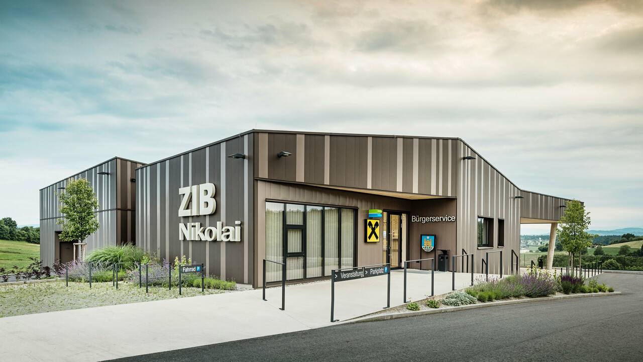 Vue de face du centre événementiel ZIB Nikolai à St. Veit, avec une façade moderne en aluminium à rayures marron-beige-gris et une grande inscription « ZIB Nikolai ». La zone d'entrée présente une signalisation claire pour les événements, le stationnement et les vélos, ainsi qu'un service citoyen intégré et une succursale de la Raiffeisenbank. En arrière-plan, des collines verdoyantes et un vaste paysage s’étendent sous un ciel nuageux.