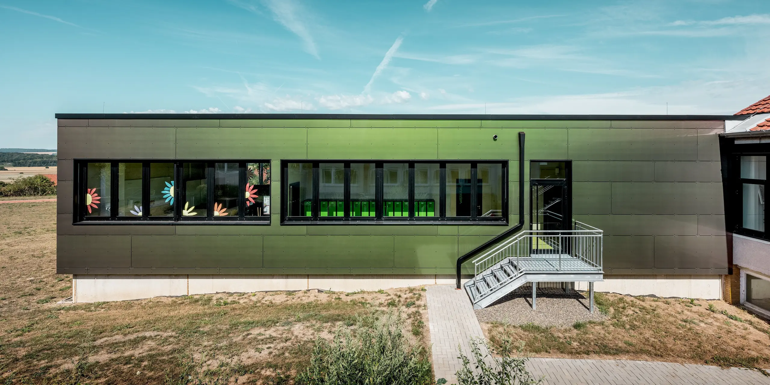 Moderner Schulanbau der Johannwolfschule Außenstelle Angerstein mit großflächiger PREFA Fassade aus Aluminium Verbundplatten in der Sonderfarbe Spring Lake. Die grünlich schimmernde Fassade umrahmt lange Fensterfronten mit bunten Blumenmotiven. Eine Außentreppe aus Metall führt zum seitlichen Eingang. Das dunkle PREFA Quadratrohr fügt sich dezent in die geradlinige Architektur ein.