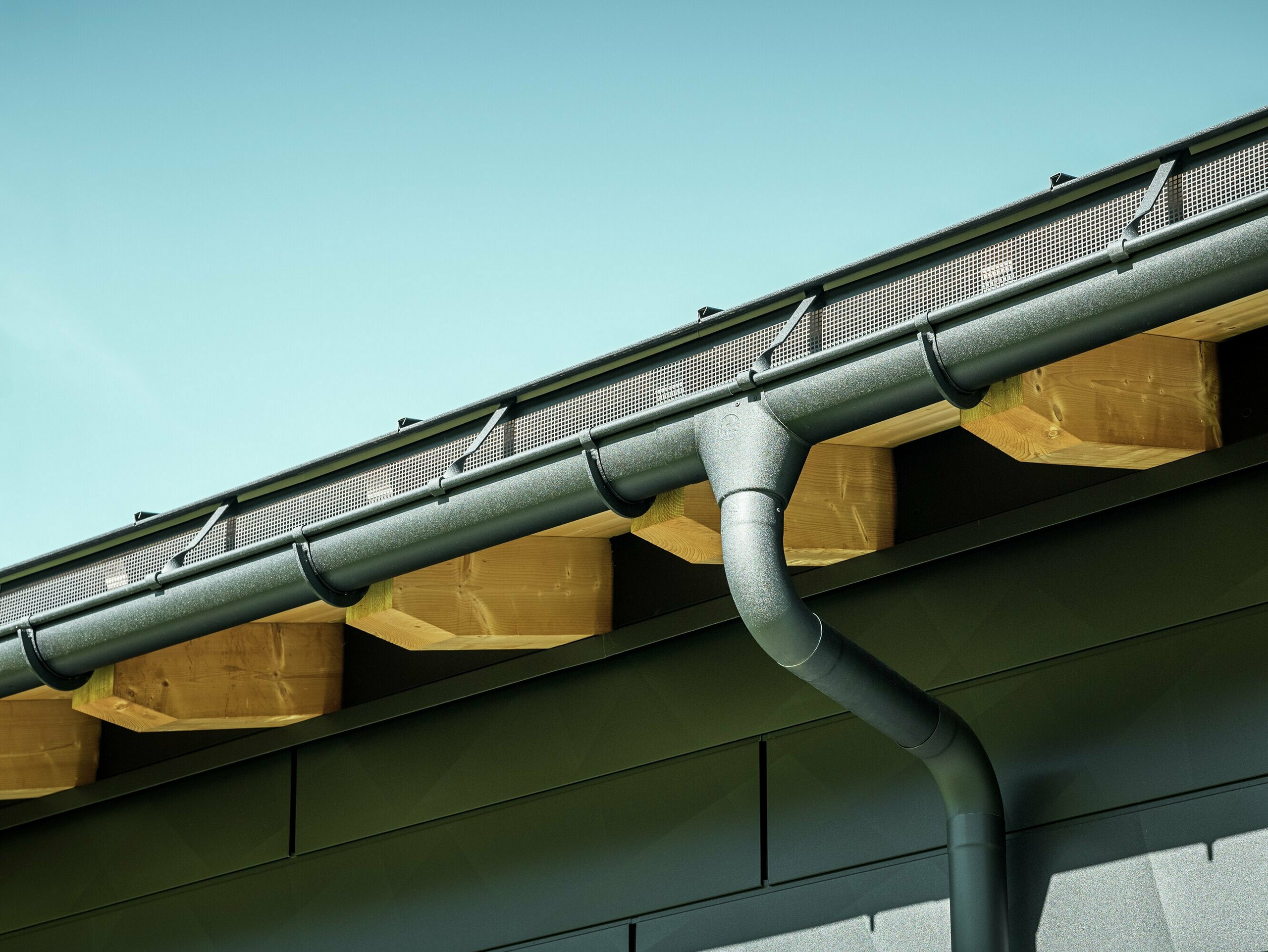 Nahaufnahme der Fassade eines modernen Einfamilienhauses in Ascharina, Schweiz. Die Gebäudeseiten wurden mit den markanten PREFA Siding.X Fassadenpaneelen in P.10 Schwarz verkleidet. Farblich abgestimmt ist die PREFA Dachentwässerung mit Hängerinne und Ablaufrohr.