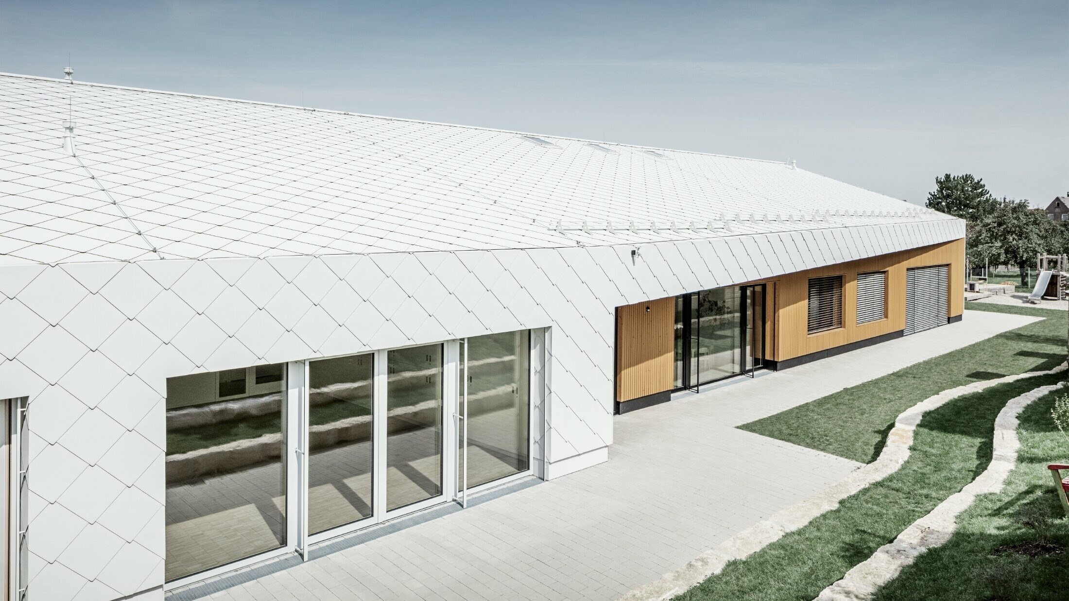 Asilo rivestito con scaglia per tetto e facciata PREFA 44 bianco PREFA, la superficie del tetto dà continuità alla facciata; ampie superfici vetrate