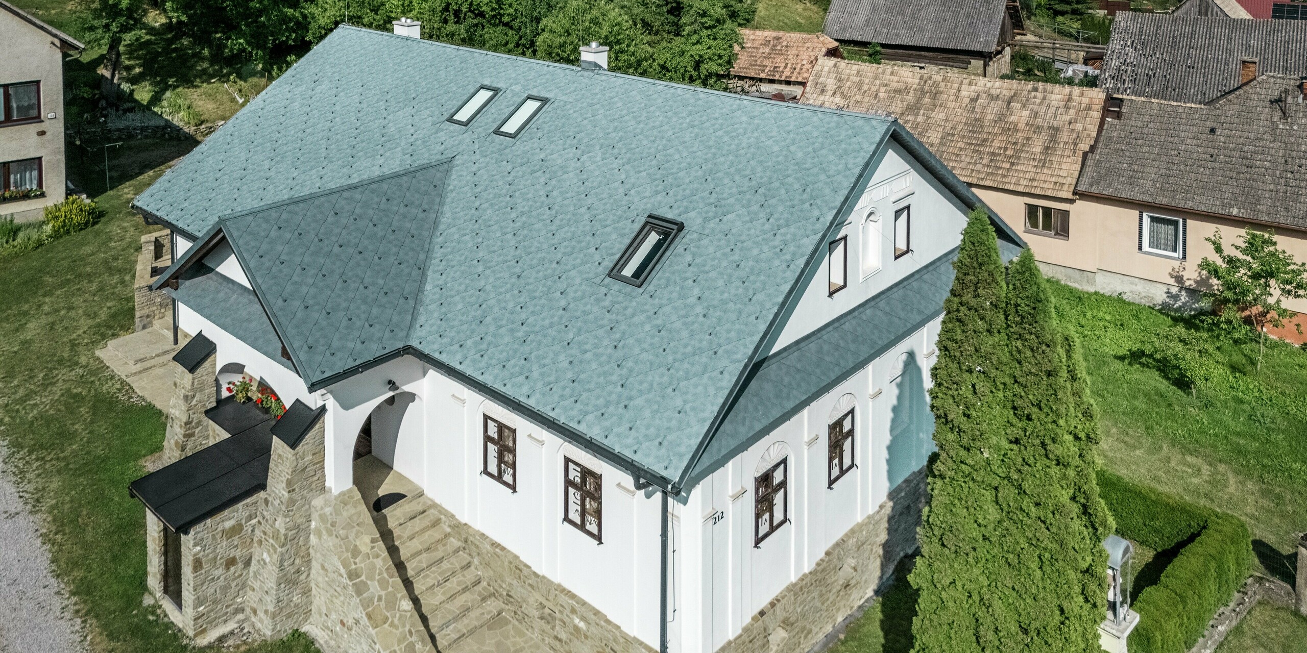 L'image présente une vue aérienne d'une maison d'artisan rénovée à Klin, en Slovaquie. Le toit à pignon simple, agrémenté d'une lucarne au-dessus de l'entrée, est recouvert de losanges PREFA 44 x 44, couleur Gris Pierre P.10. La surface du toit, caractérisée par des losanges symétriques, confère au bâtiment une allure à la fois moderne et élégante. Les puits de lumière intégrés apportent un éclairage naturel supplémentaire et soulignent la fonctionnalité de la conception. La toiture en aluminium offre de nombreux avantages : légère, durable et résistante à la corrosion, elle est idéale pour la rénovation de bâtiments historiques, car elle minimise la charge sur le toit tout en offrant une excellente résistance aux intempéries.