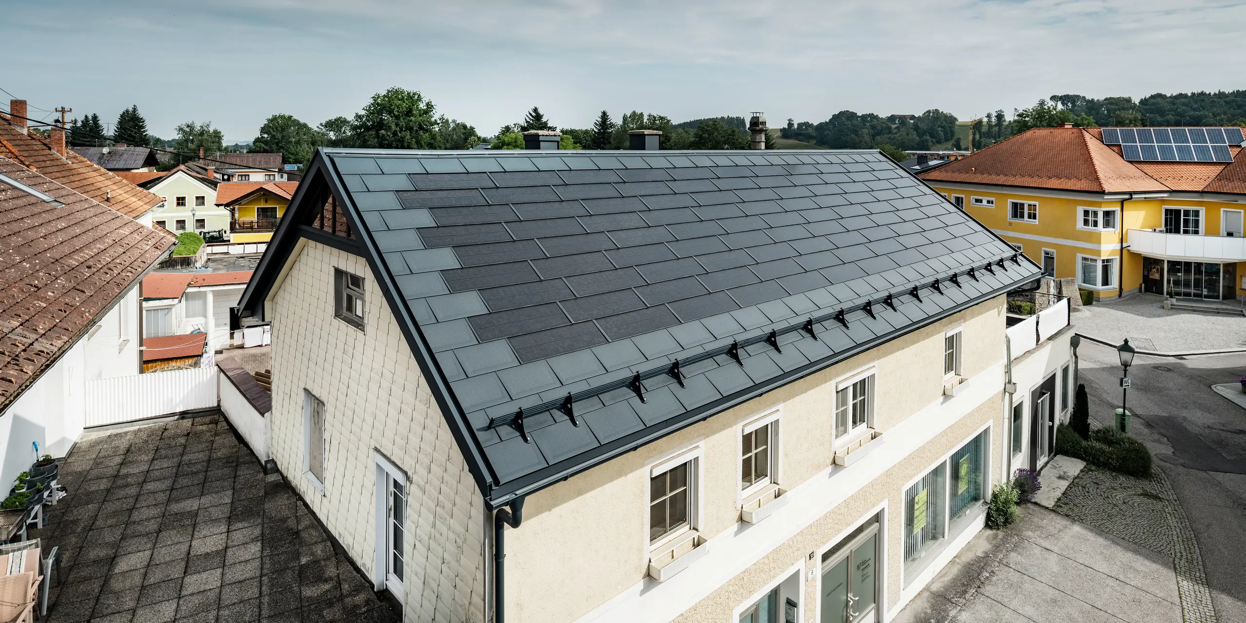 Blick von oben auf ein Einfamilienhaus in Mettmach, Österreich. Das ältere Gebäude wurde mit anthrazitfarbenen PREFA Solardachplatten saniert. Die Schneefangsysteme und die klare Linienführung der Solardachplatten unterstreichen die saubere Verarbeitung der PREFA Aluminiumprodukte. Im Hintergrund kontrastiert das orangefarbene Ziegeldach eines Nachbargebäudes. Auf dem Dach des benachbarten Einfamilienhauses sind Photovoltaikmodule in einer klassischen Aufdach-Lösung installiert, was die Vielfalt der energieeffizienten Dachgestaltung zeigt.