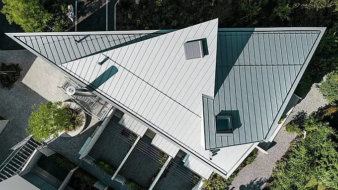 L'immagine mostra il tetto di un moderno edificio a più piani a Zollikerberg, in Svizzera. Il tetto è rivestito con Prefalz in patina grigio, un sistema di copertura in alluminio durevole e resistente alla corrosione prodotto da Prefa. La struttura del tetto è inclinata e dal design geometrico, con diverse finestre e un aspetto elegante e moderno. Intorno all'edificio sono visibili aree verdi ben curate e marciapiedi.