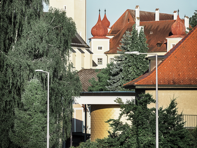 Aufnahme von Buchkirchen mit der Landesmusikschule und anderen Gebäuden und Bäumen im Hintergrund. Die Fassade wurde mit PREFA Wandschindeln in der Sonderfarbe Mayagold verkleidet und blitzt durch Bäume und Gebäude hervor.