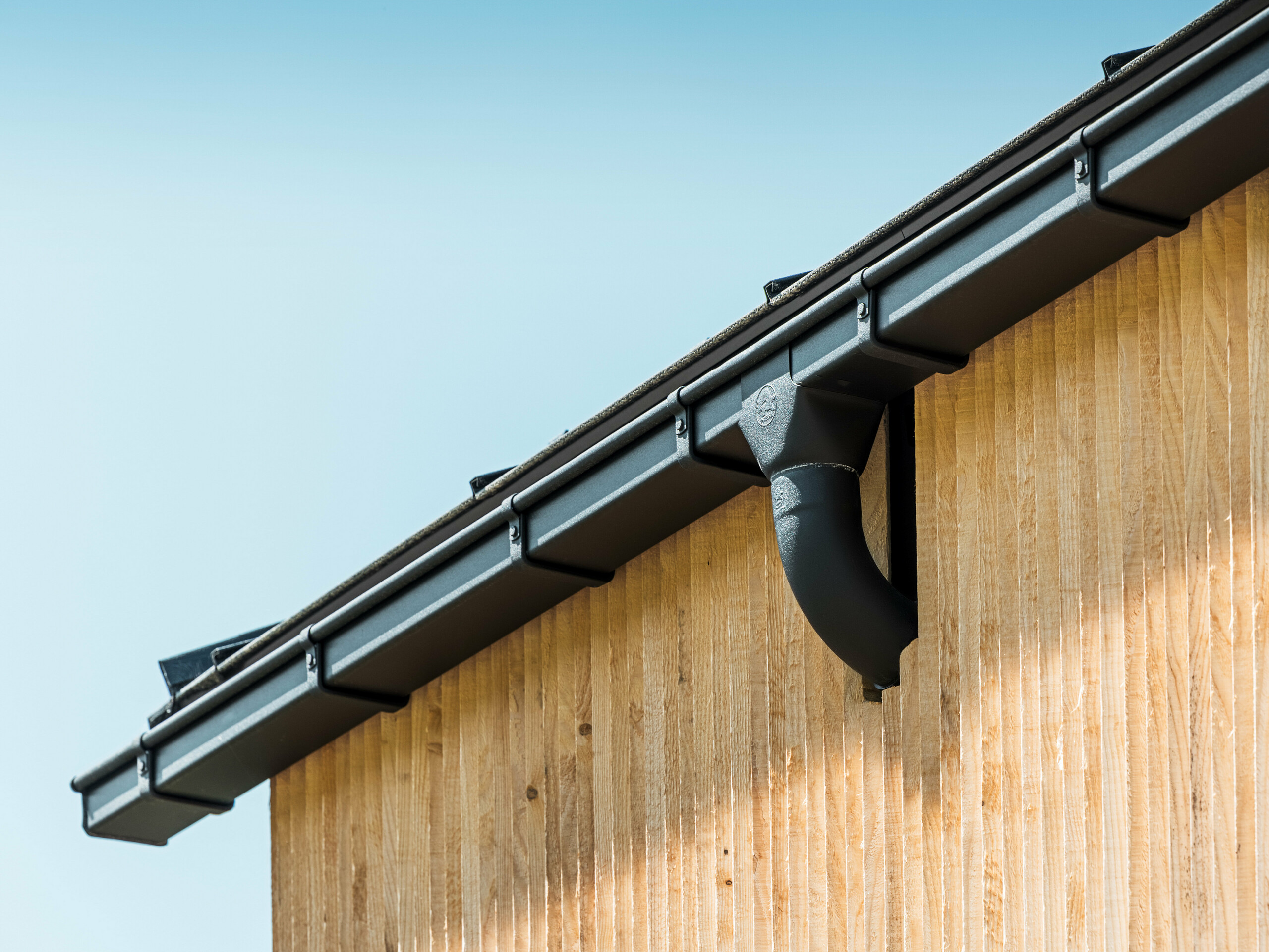 Angolo dell'edificio con canale quadro PREFA in nero, con cassetta di raccolta, fissato con cicogne per canali di gronda PREFA, la facciata è rivestita in legno.