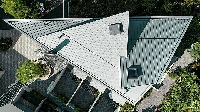 L'immagine mostra il tetto di un moderno edificio a più piani a Zollikerberg, in Svizzera. Il tetto è rivestito con Prefalz in patina grigio, un sistema di copertura in alluminio durevole e resistente alla corrosione prodotto da Prefa. La struttura del tetto è inclinata e dal design geometrico, con diverse finestre e un aspetto elegante e moderno. Intorno all'edificio sono visibili aree verdi ben curate e marciapiedi.