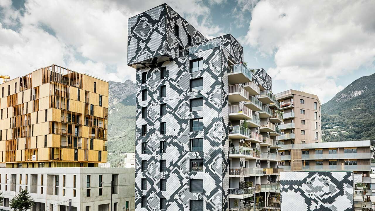 modernes und neues Wohnhaus in Grenoble, mit einer Aluminium Wandraute verkleidet, das Verlegemuster entspricht der Schlangenhaut einer Python in anthrazit, hellgrau, naturblank und silbermetallic, daneben ein Sandbrauner Wohnblock vor bewölktem Himmel