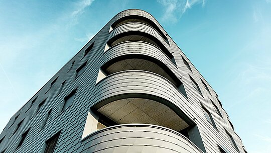 Dynamische Untersicht eines modernen Wohn- und Geschäftshauses mit markant gerundeter Ecke und fließenden Balkonbändern. Die schuppenartige Fassade aus PREFA Wandschindeln in P.10 Hellgrau legt sich wie eine präzise verarbeitete Aluminiumhaut über das Gebäude und betont die weichen Radien. Die homogene Oberfläche unterstreicht die klare Linienführung, während das leichte, witterungsbeständige PREFA Aluminium selbst bei starken Rundungen höchste Formstabilität, Langlebigkeit und Wartungsarmut garantiert – ein architektonischer Eyecatcher vor strahlend blauem Himmel.