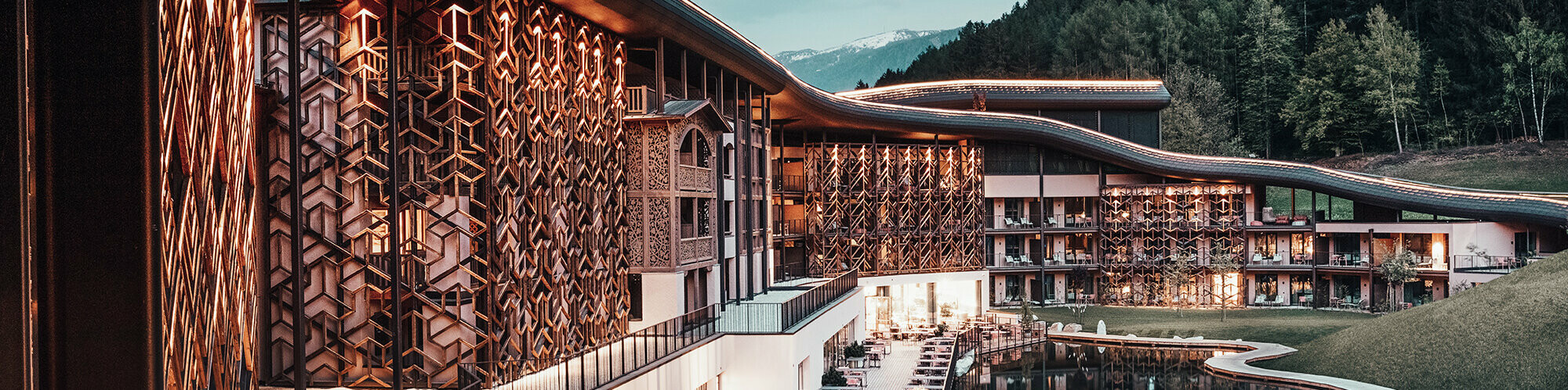 Abendaufnahme, warmes Licht beleuchtet die Holzfassade und das Aluminiumdach, unten die Terrasse mit Sitzgelegenheiten, davor der See, im Hintergrund der dunkelgrüne Wald