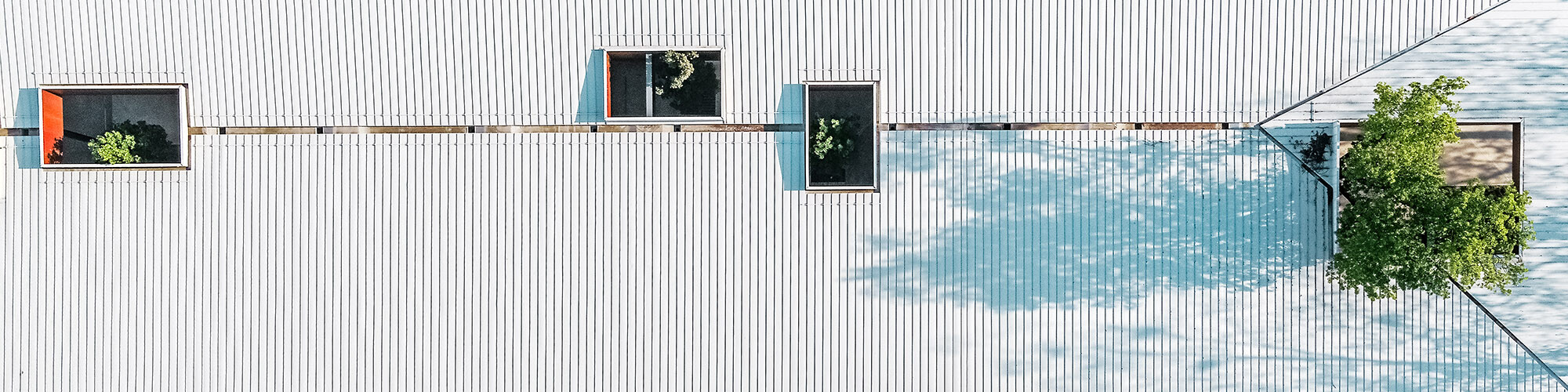 La photo montre une vue aérienne du toit Prefalz blanc du hall de la gare routière « Autobusni Kolodvor Slavonski Brod » en Croatie. Le toit est percé de plusieurs ouvertures rectangulaires à travers lesquelles poussent les arbres verts, créant un lien harmonieux entre l'architecture et la nature. Les lignes épurées et la surface blanche du toit, brisées par l'ombre des arbres, forment un motif intéressant. La photo met en évidence le design innovant et écologique du bâtiment, qui se distingue par sa fonctionnalité et son intégration esthétique dans l'environnement.