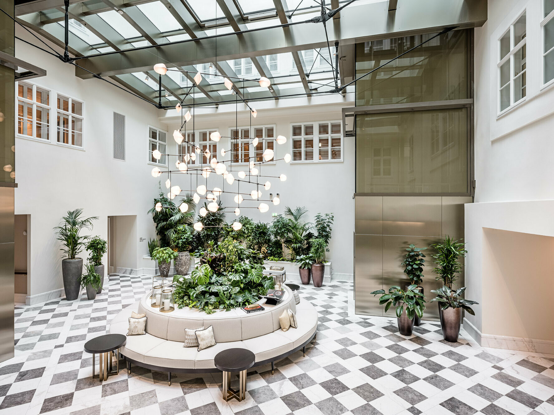Intérieur de l’hôtel de luxe, où prédominent les couleurs bronze, gris et blanc : des fauteuils et de grandes plantes en pot sont disposés sur un sol en marbre à damier, un éclairage moderne est suspendu au plafond de la pièce. 