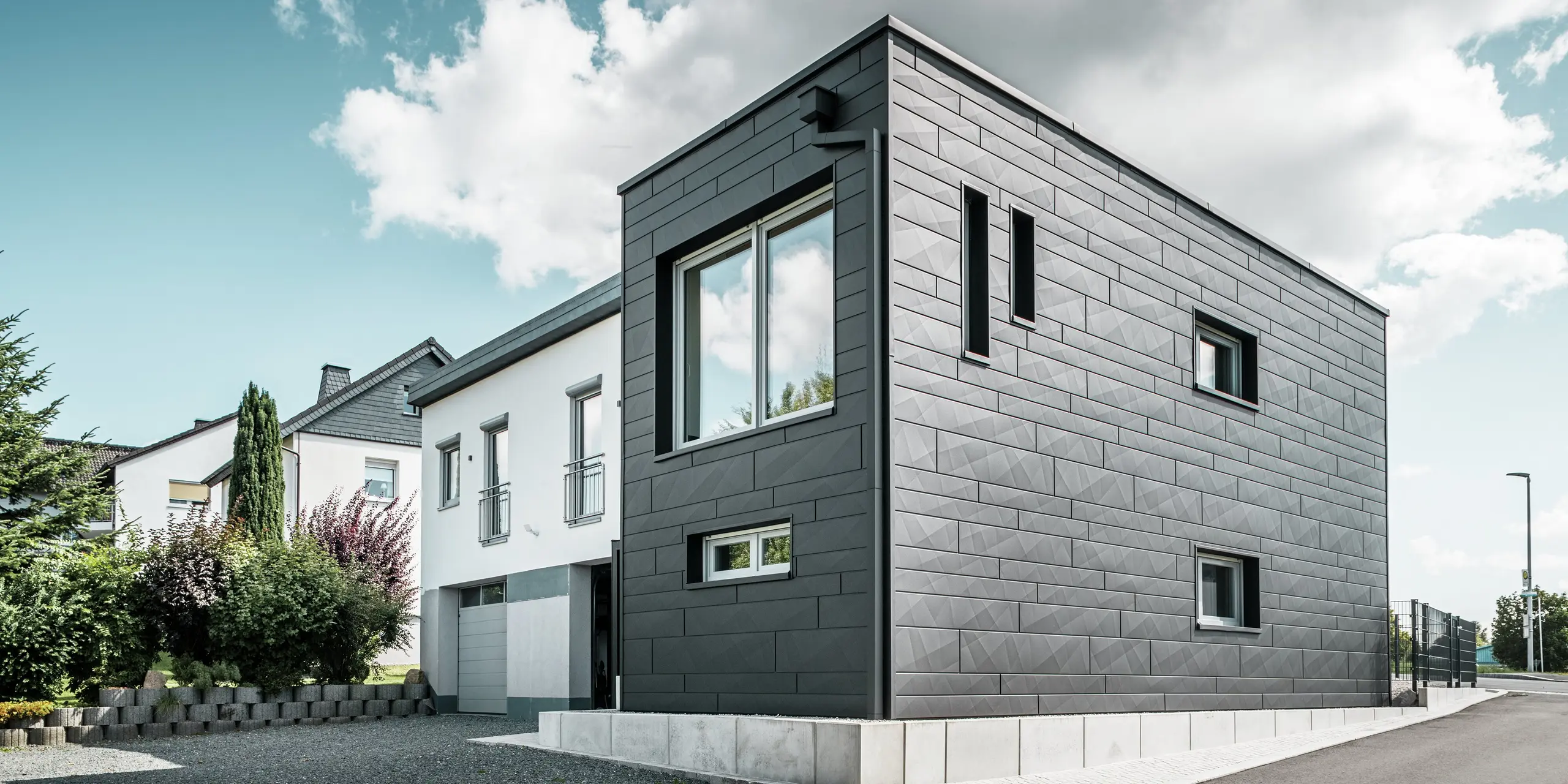 Die stilvolle und moderne Fassade des sanierten Einfamilienhauses in Teuschnitz zeigt die klar strukturierten PREFA Siding.X Paneele in P.10 Schwarz. Die geometrische Anordnung der hochwertigen Blechpaneele verleiht dem Gebäude eine markante, zeitlose Ästhetik. Die Fenster sind von schwarzen Aluminium Verbundplatten umrahmt, was das Design harmonisch abrundet. Die integrierte PREFA Dachentwässerung mit dem Quadratrohr sorgt für eine funktionale und ästhetische Lösung.