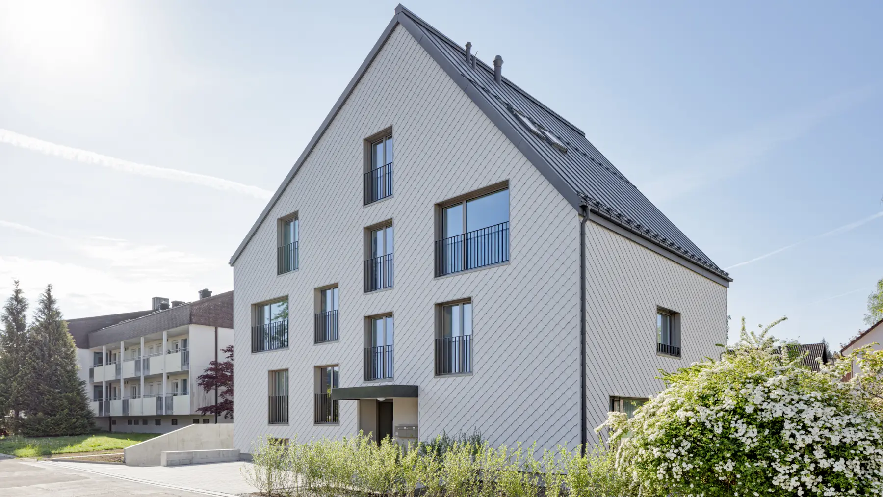 Das Bild zeigt ein modernes Mehrfamilienhaus mit einer eleganten Fassade, die mit PREFA Wandrauten 20 × 20 in Prefaweiß verkleidet ist. Das Gebäude hat ein schräges, steiles Dach, das mit einem PREFALZ-Dach in der Farbe P.10 Dunkelgrau gedeckt ist. Die Kastenrinne ebenso in P.10 Dunkelgrau fügt sich harmonisch in das Design ein. Der Außenbereich des Hauses ist von Grünflächen umgeben.