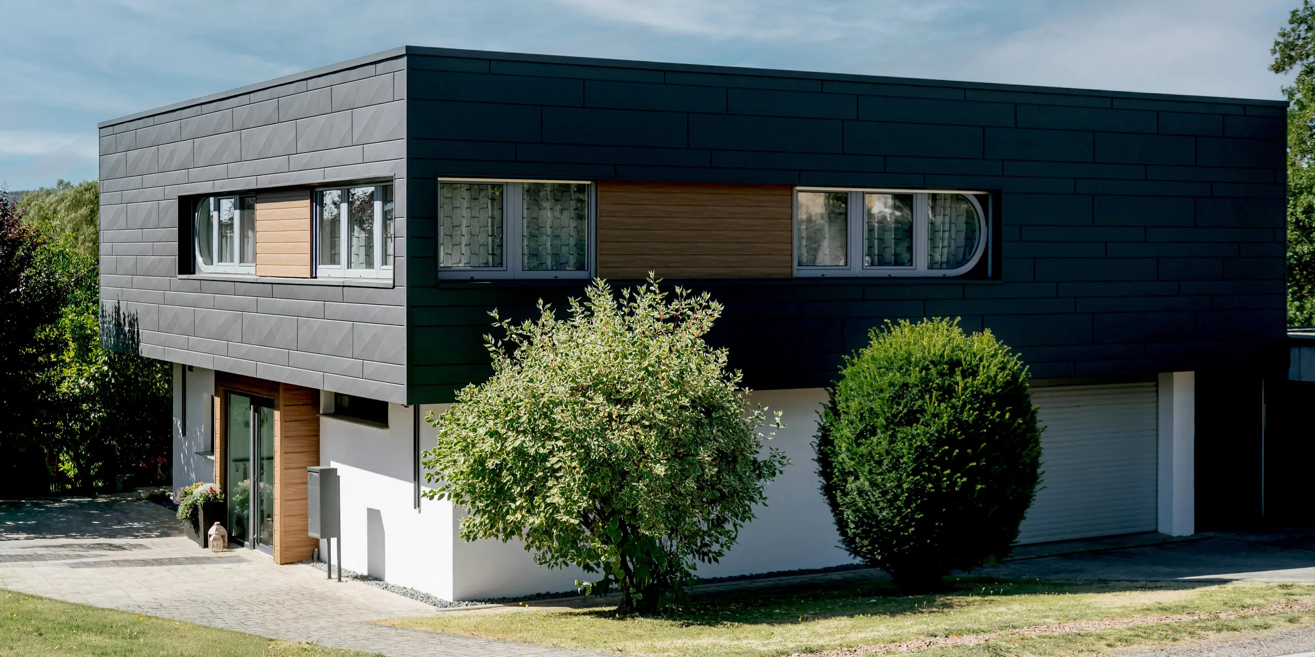 Großzügige Ansicht eines modernen Einfamilienhauses in Marksuhl mit prägnanter PREFA Aluminiumfassade. Die horizontal verlegten PREFA Siding.X Paneele in P.10 Dunkelgrau verleihen dem Baukörper eine klare, puristische Form. Akzentflächen aus PREFA Siding in Walnuss braun schaffen natürliche Wärme. Die langlebige Aluminiumverkleidung bietet zuverlässigen Schutz vor Witterung und überzeugt durch hochwertige Ästhetik.