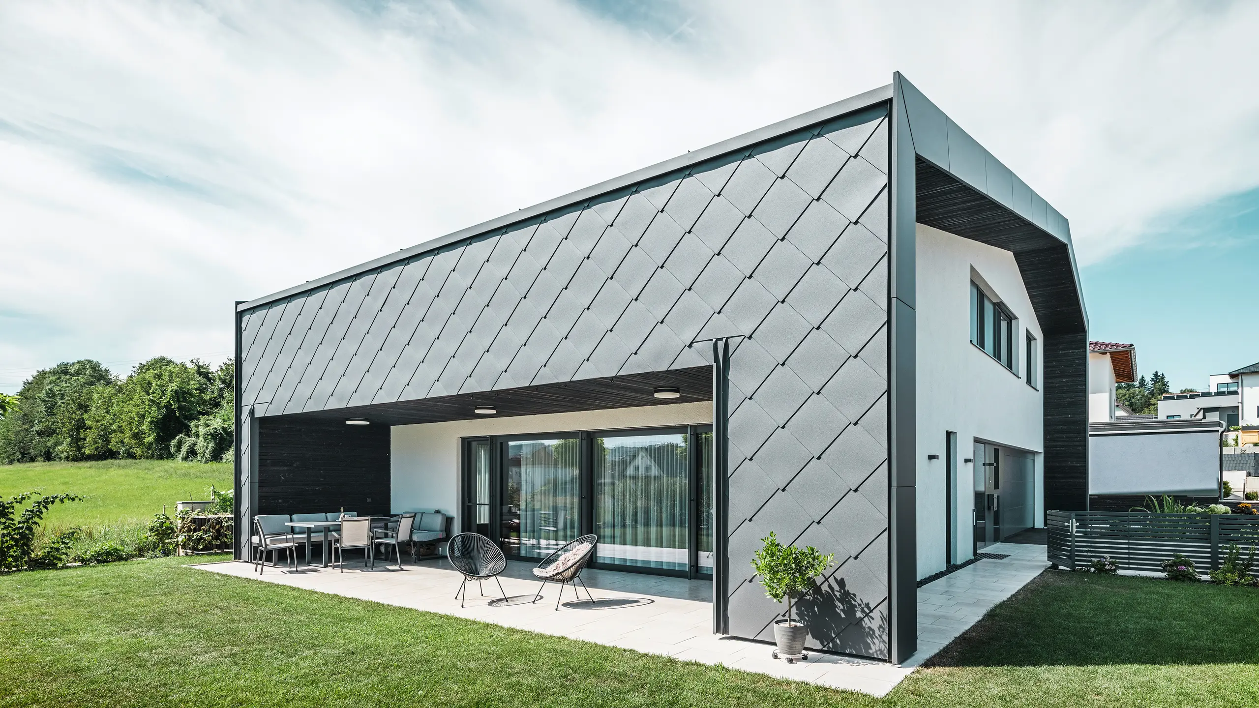 Giardino ben curato di una casa unifamiliare ad Aschach. L'edificio è elegantemente rivestito con scaglie per facciata PREFA in alluminio grigio scuro 44 × 44. La terrazza coperta offre un accogliente posto a sedere con vista sul prato verde e rigoglioso. L'elegante rivestimento in alluminio, in combinazione con le superfici intonacate bianche, garantisce un'architettura moderna e resistente alle intemperie. Le grandi superfici vetrate lasciano entrare molta luce naturale all'interno e creano un'atmosfera aperta e luminosa.