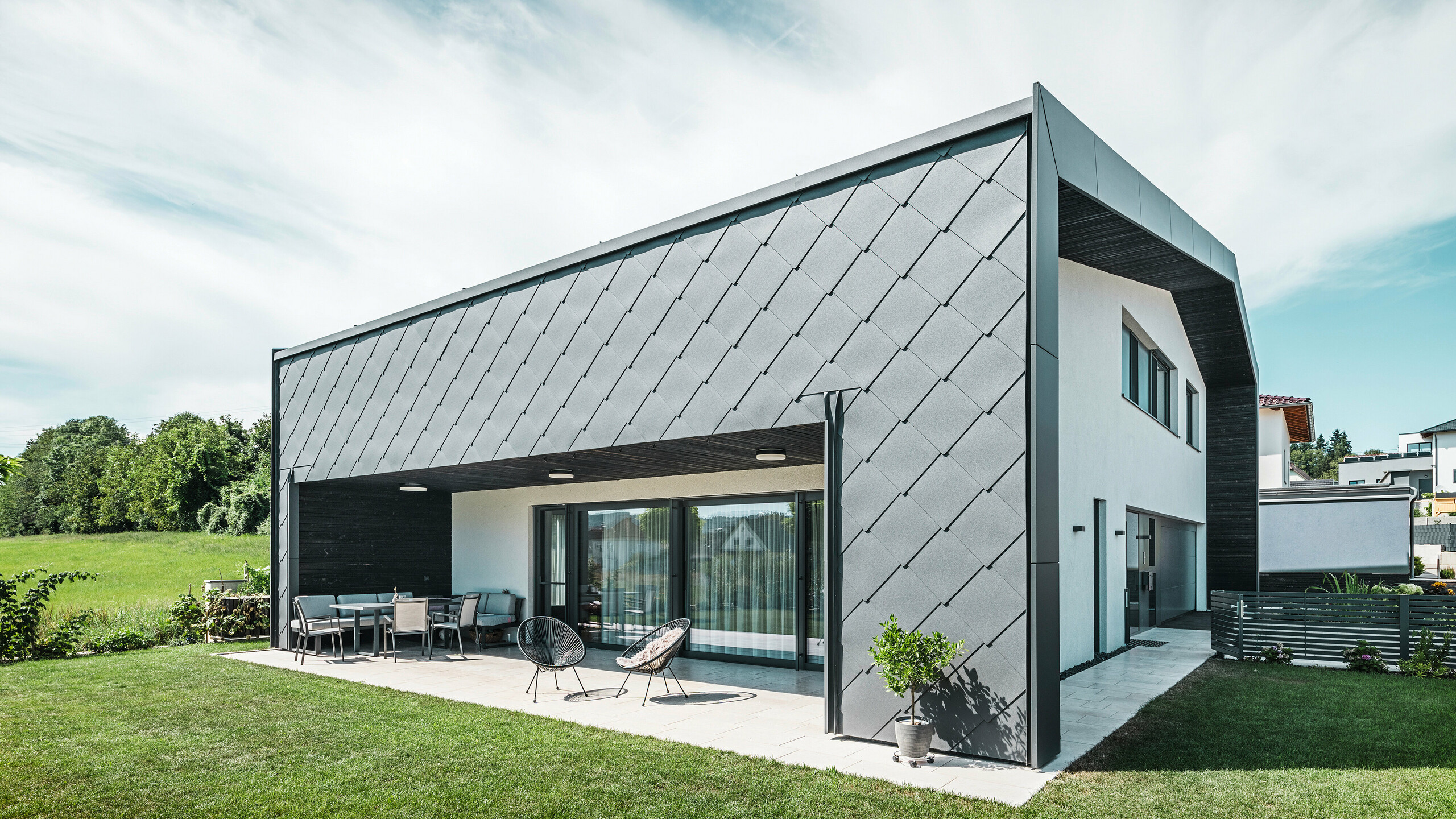 Gepflegter Gartenbereich eines Einfamilienhauses in Aschach. Das Gebäude ist stilvoll eingedeckt mit dunkelgrauen PREFA Aluminium-Wandrauten 44 × 44. Die überdachte Terrasse bietet einen gemütlichen Sitzplatz mit Blick auf die saftig grüne Rasenfläche. Die elegante Aluminiumverkleidung in Kombination mit weißen Putzflächen sorgt für eine moderne und witterungsbeständige Architektur. Große Fensterflächen lassen viel Tageslicht in den Innenraum und schaffen eine offene, lichtdurchflutete Atmosphäre.