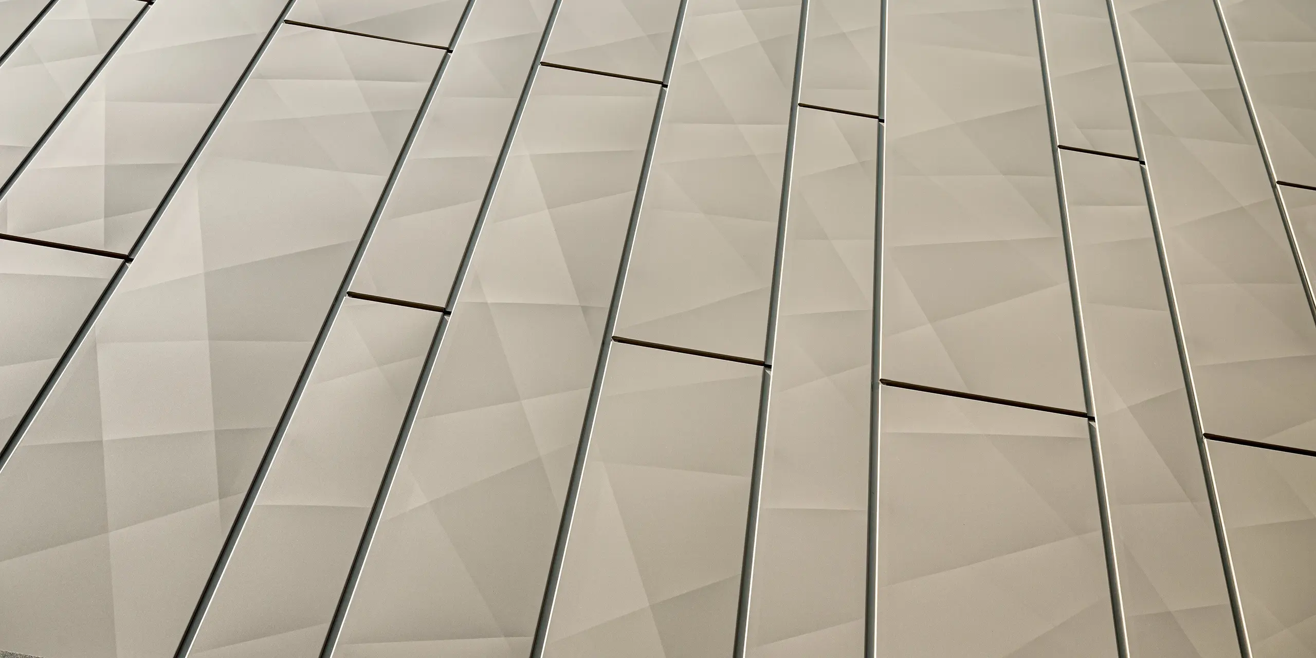 L'image présente un gros plan des panneaux Siding.X sur la façade en aluminium du centre commercial de Rohrbach. Ces éléments de façade, à la finition bronze élégante, arborent une structure géométrique qui crée une texture tridimensionnelle dynamique grâce à des lignes précises et des plis discrets. La fluidité des transitions et la finesse du découpage des panneaux confèrent à la façade une allure moderne et raffinée. Outre leur esthétique, ces éléments de façade sont extrêmement résistants aux intempéries, ce qui garantit un entretien facile et une longue durée de vie.