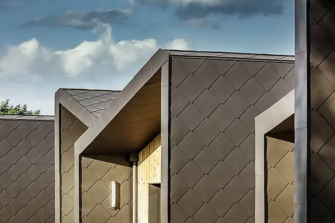 L'immagine mostra una facciata moderna rivestita con le scaglie PREFA per tetto e facciata nel colore marrone testa di moro. La struttura a rombo del rivestimento in alluminio enfatizza le linee chiare e offre una soluzione durevole e resistente alle intemperie per i sistemi di copertura e facciata. La combinazione tra i diamanti di metallo scuro e i caldi elementi in legno conferisce all'edificio un aspetto elegante e senza tempo. Questa architettura moderna e di alta qualità è ideale per progetti edilizi efficienti dal punto di vista energetico ed esteticamente gradevoli.