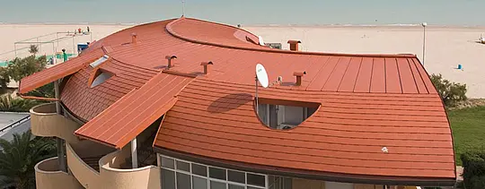 Gerundetes Dach einer Wohnhausanlage, eingedeckt mit der PREFA Dachschindel sowie Stehfalzdach - Prefalz - von PREFA in Ziegelrot.