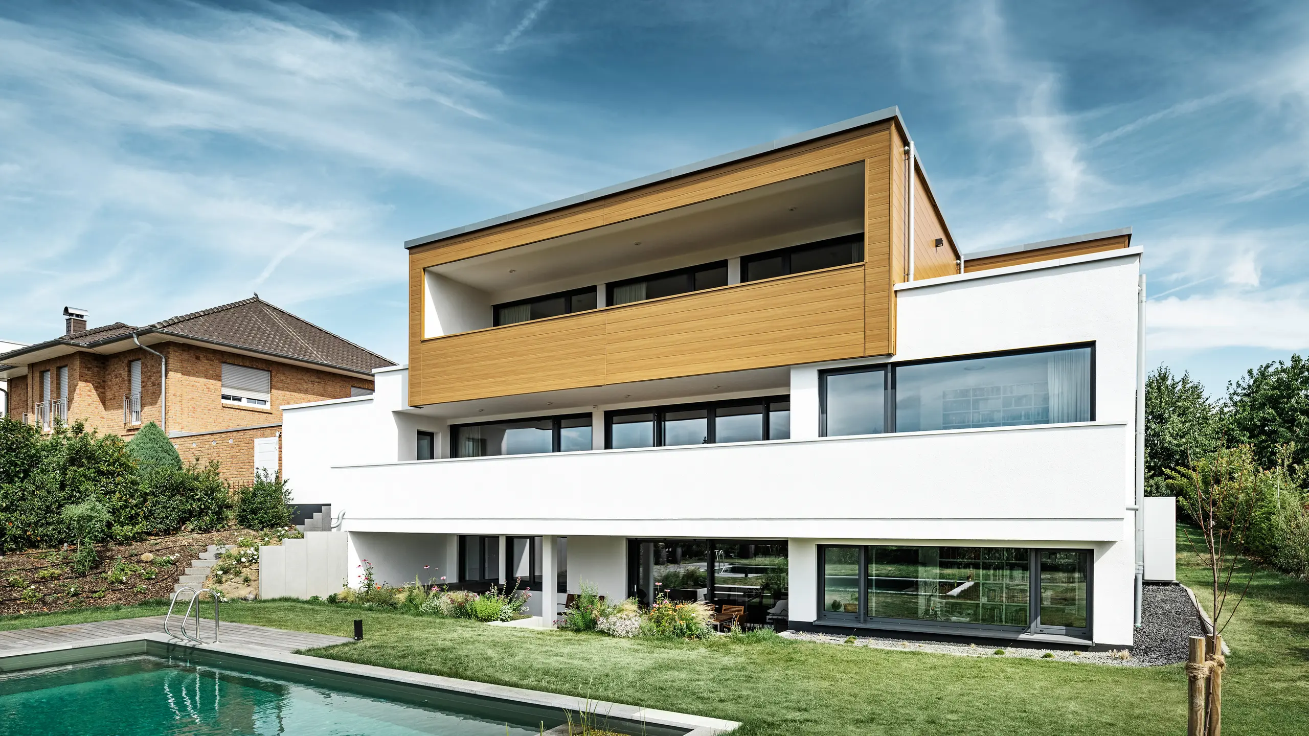 Il retro di una moderna casa indipendente a Usingen, in Germania. Il prato verde, la piscina e un giovane albero da giardino sono visibili nella parte inferiore dell'immagine. L'accattivante casa ha una base bianca costituita da una facciata intonacata, una facciata in alluminio al piano superiore realizzata con doga PREFA in rovere naturale e ampie finestre con telaio color antracite. Da lontano, la casa vicina con facciata in mattoni è visibile sul bordo sinistro dell'immagine.