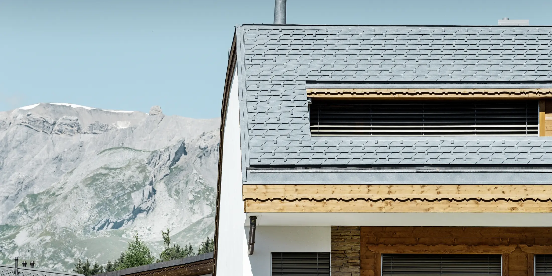 Mehrfamilienhaus in Crans-Montana mit Bergen im Hintergrund und einer Fassade mit verspielten Holzelementen mit einem PREFA Aluminium Dachschindel-Dach in steingrau 