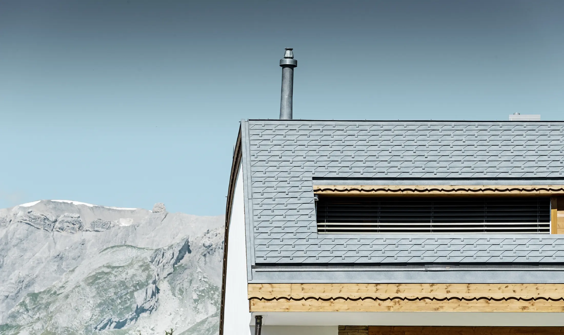 Casa plurifamiliare in Crans-Montana con montagne sullo sfondo e una facciata con elementi decorativi in legno e con una copertura in scandole PREFA grigio pietra