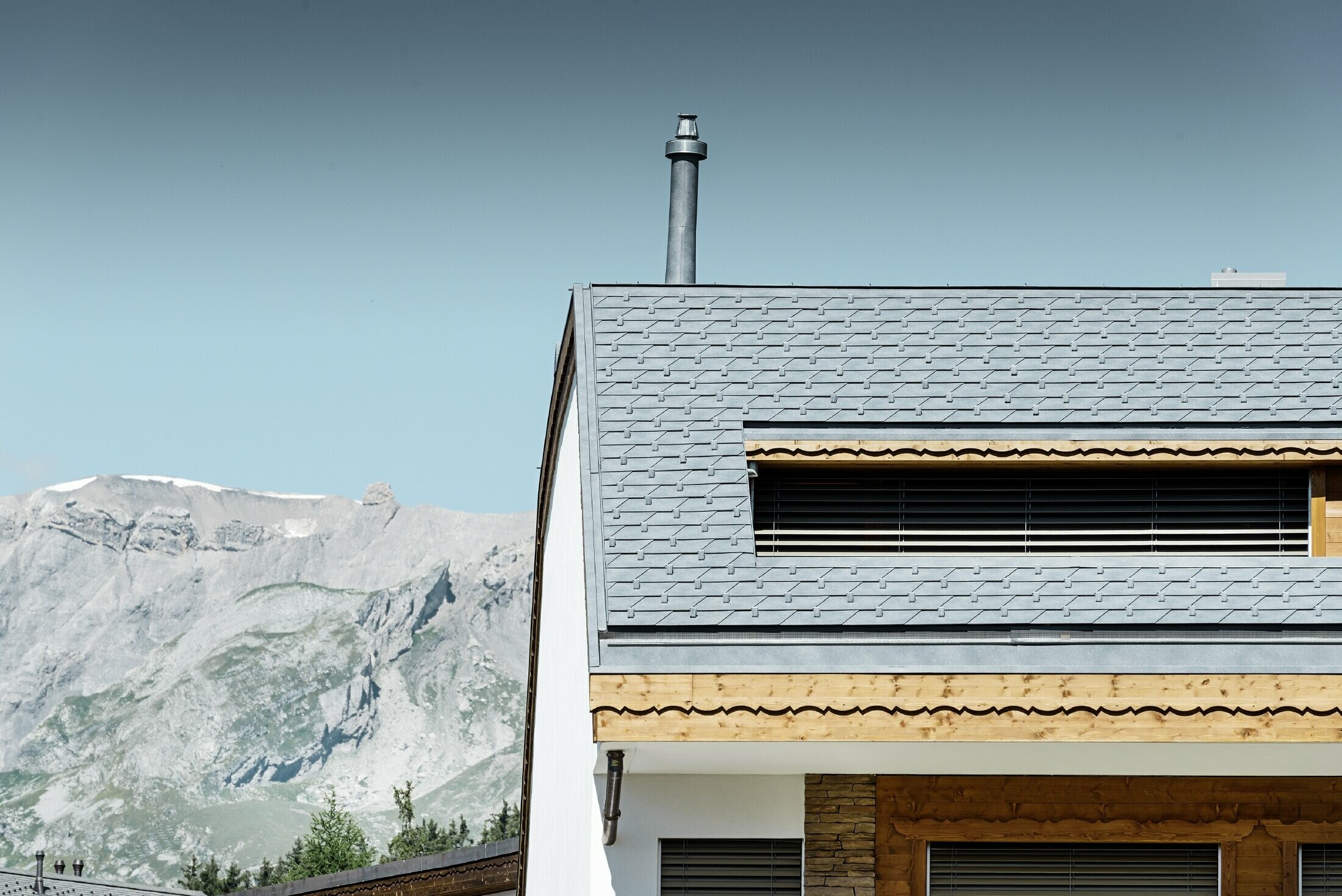 Casa plurifamiliare in Crans-Montana con montagne sullo sfondo e una facciata con elementi decorativi in legno e con una copertura in scandole PREFA grigio pietra