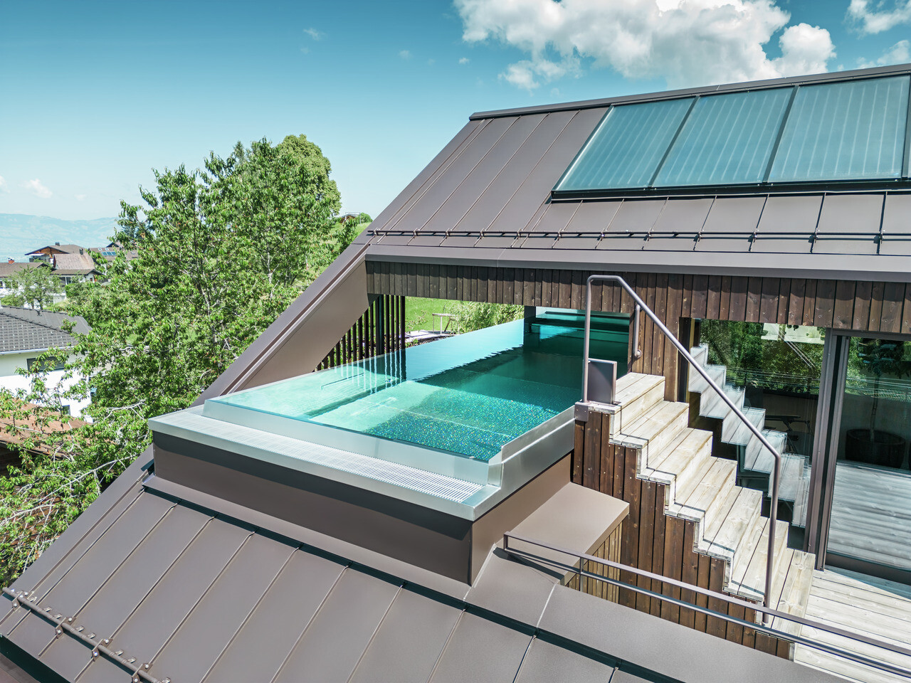 Vue aérienne d'une maison moderne avec un toit en Prefalz PREFA brun noisette. Un toit-terrasse avec piscine offre un espace de détente exclusif avec vue sur le paysage vallonné et verdoyant.