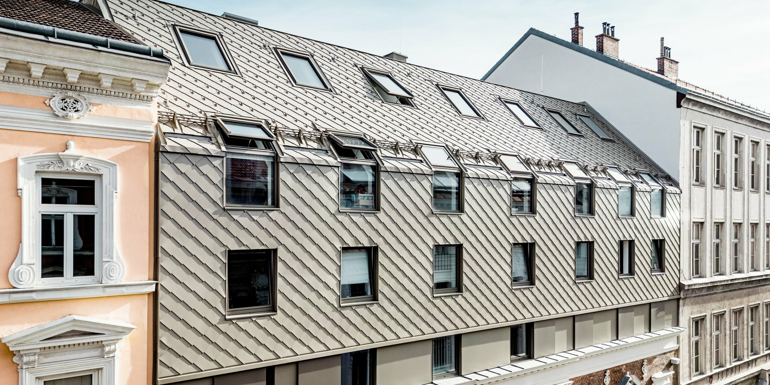 Das Bild zeigt die prägnante Erweiterung eines Wiener Wohngebäudes, bei dem moderne PREFA Rauten 29×29 in Bronze die obere Etage zieren. Die Rauten verleihen der Architektur eine elegante und zeitgemäße Note, die perfekt mit der historischen Fassade darunter harmoniert. Besonders auffällig sind die Dachgauben, die das Dach beleben und eine interessante Struktur schaffen. Der Kontrast zwischen der klassischen Ziegelmauer und den modernen Aluminium-Rauten betont die gelungene Mischung aus Tradition und Innovation in der Wiener Architektur.