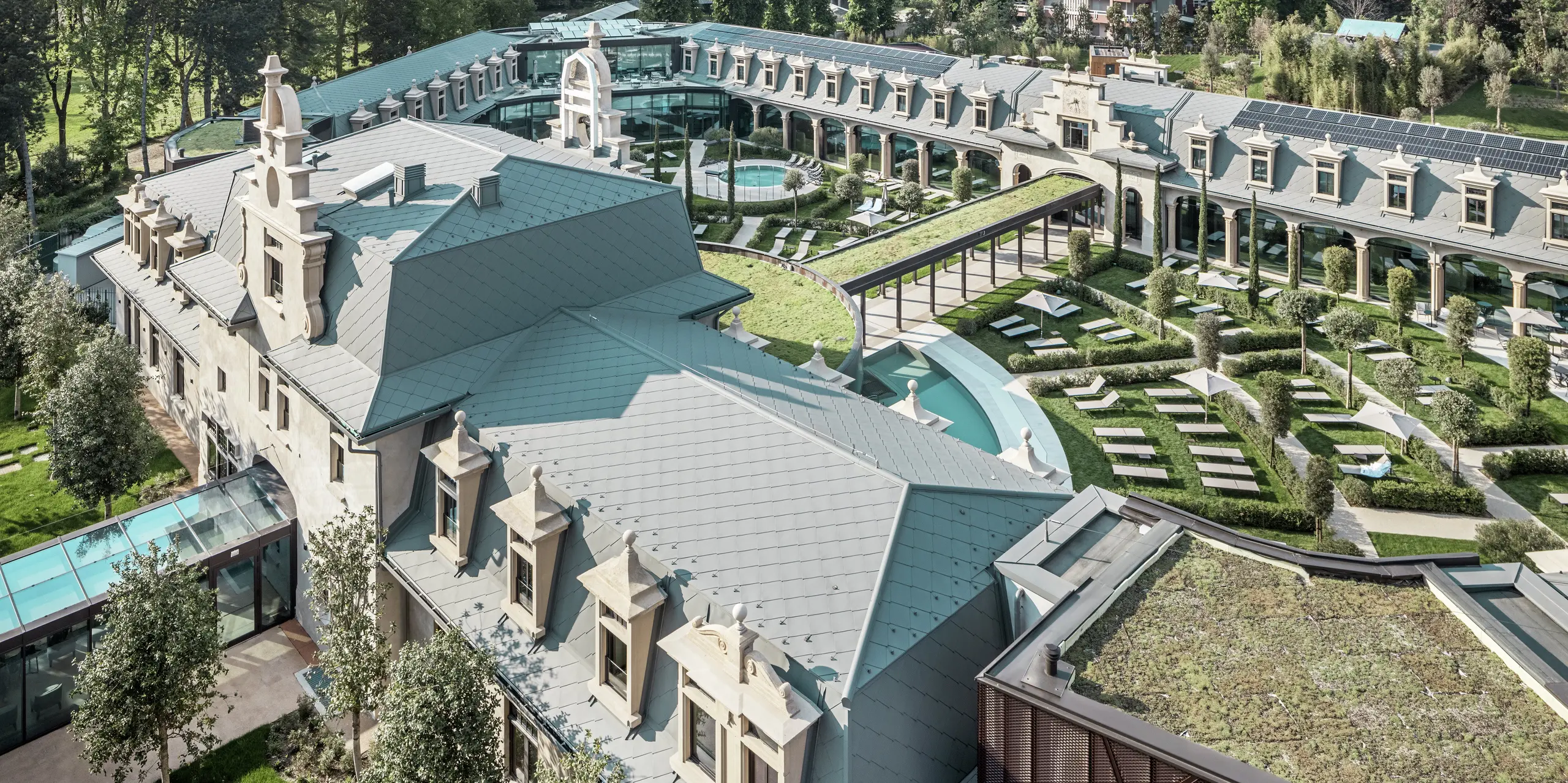 [Translate to Italiano:] Panoramablick auf den vollständig sanierten Thermenkomplex „De Montel – Terme Milano“ mit großzügiger Gartenanlage, geschwungenen Wegen und Liegebereichen rund um zahlreiche Thermalbecken. Die Dächer sind durchgängig mit der PREFA Dachraute 44 × 44 in P.10 Hellgrau eingedeckt, ergänzt durch nahtlos integrierte Photovoltaikmodule und begrünte Flächen. Historische Architektur und nachhaltige Materialien verbinden sich zu einem innovativen, urbanen Erholungsraum.