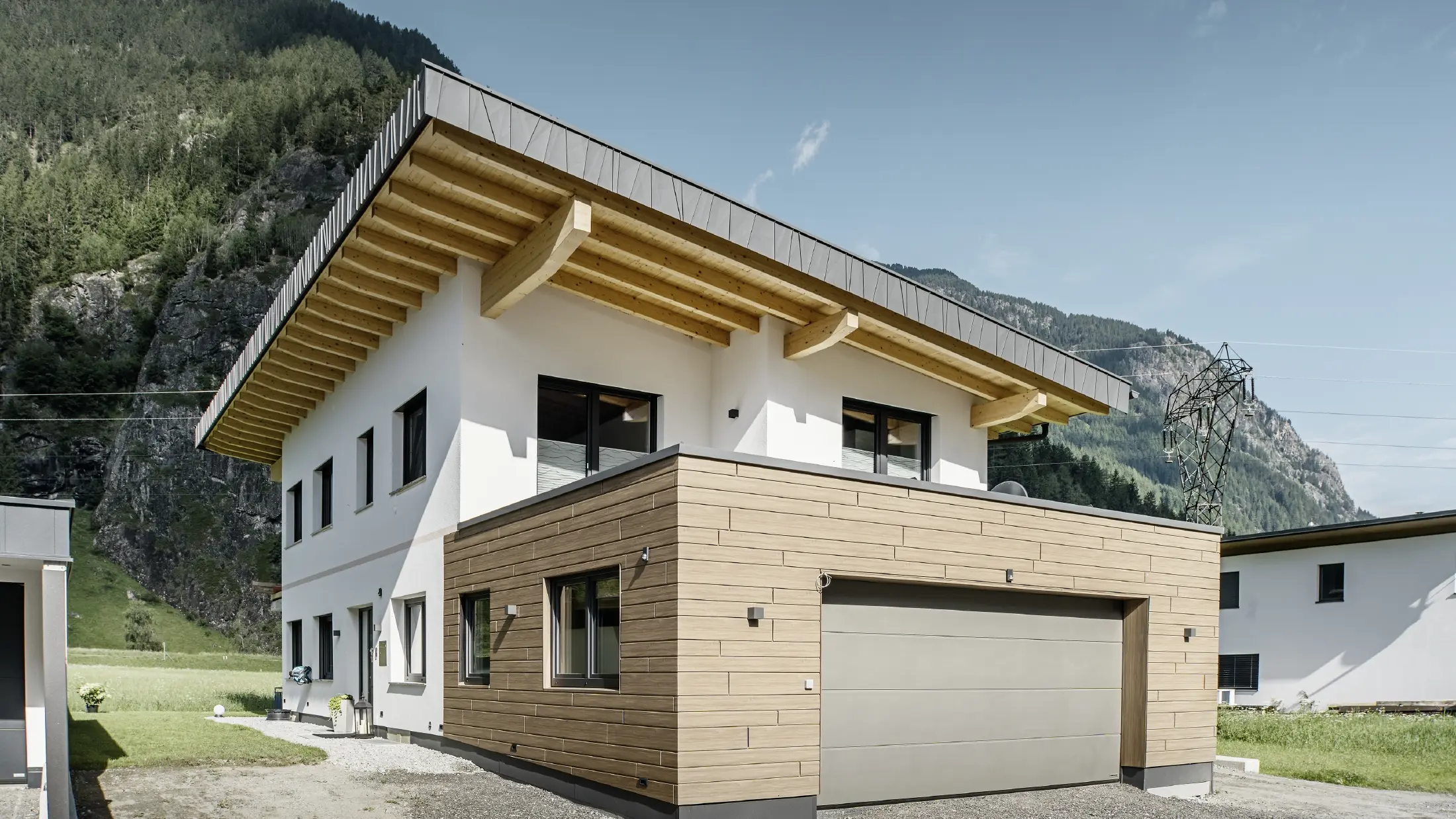 Moderna casa unifamiliare con facciata ristrutturata: facciata esterna rinnovata con rivestimento orizzontale effetto legno, abbinato a superfici intonacate di bianco, rivestimento del garage integrato e architettura contemporanea in un contesto alpino.