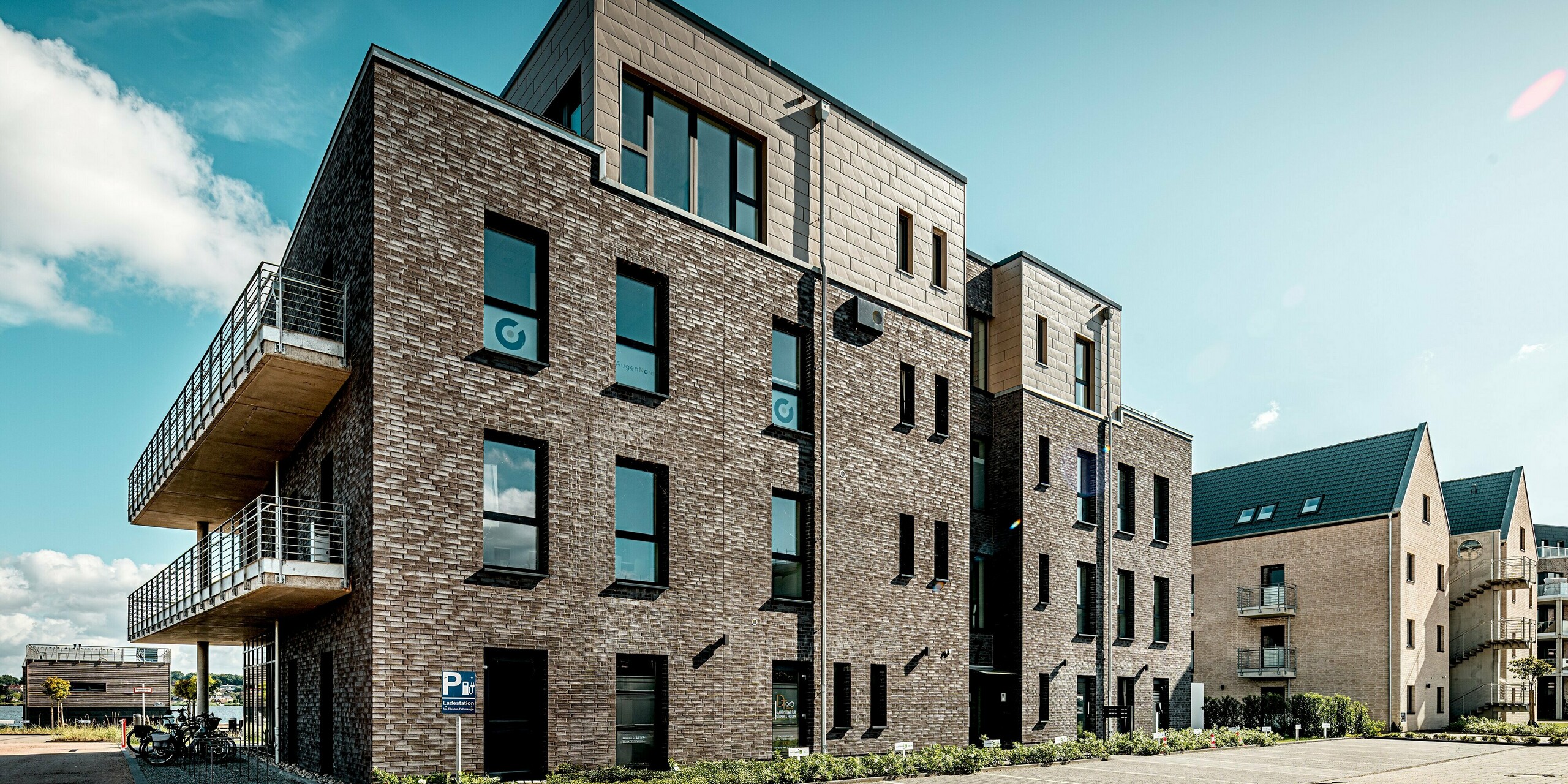 Das Bild zeigt ein Gebäude der Wohnanlage „Hafen zur Freiheit“, das durch die Kombination aus dunkler Klinkerfassade und den markanten PREFA Siding.X Paneelen in Bronze im Staffelgeschoss besonders modern wirkt. Die klar strukturierten Aluminiumflächen setzen einen eleganten Kontrast zur traditionellen Ziegeloptik und verleihen dem Baukörper eine dynamische, hochwertige Ausstrahlung. Großzügige Fenster und seitliche Balkonflächen betonen die offene Architektur. Dank der langlebigen, witterungsfesten PREFA Elemente bleibt die Fassade auch in der maritimen Umgebung dauerhaft geschützt und optisch beständig.