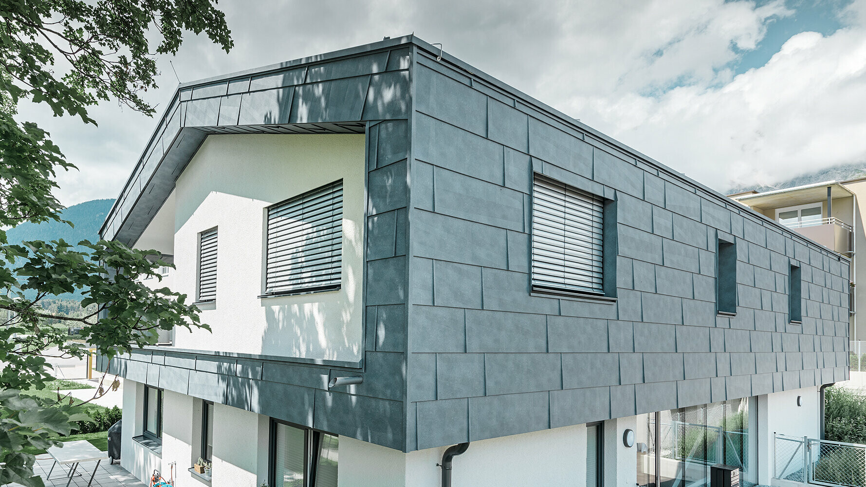 Maison individuelle moderne sans avancée de toit. L’étage supérieur a été recouvert de panneaux de façade FX.12 PREFA couleur gris pierre et le reste de la façade a été crépi en blanc.