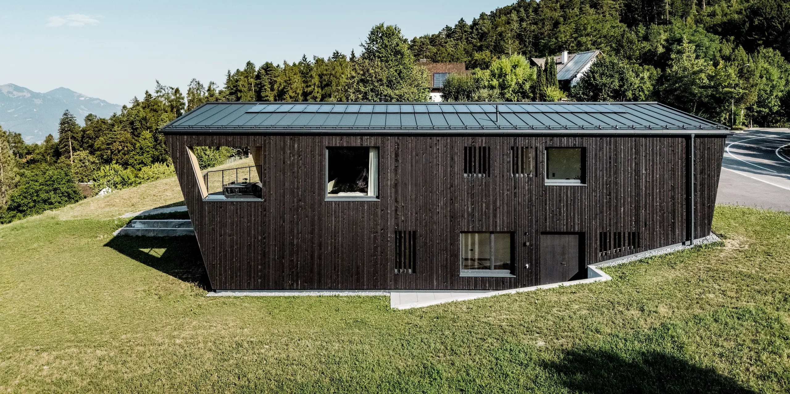 Seitenansicht des modernen Einfamilienhauses in Röthis: Das PREFALZ Satteldach in P.10 Anthrazit mit integrierten Solarmodulen PREFALZ setzt einen eleganten Akzent über der dunklen Holzfassade. Die markante Gebäudeform mit geschütztem Balkon fügt sich harmonisch in die grüne Hanglage ein. Die langlebige, leichte Aluminiumdeckung bietet zuverlässigen Witterungsschutz und verbindet zeitgemäßes Design mit nachhaltiger Energiegewinnung.
