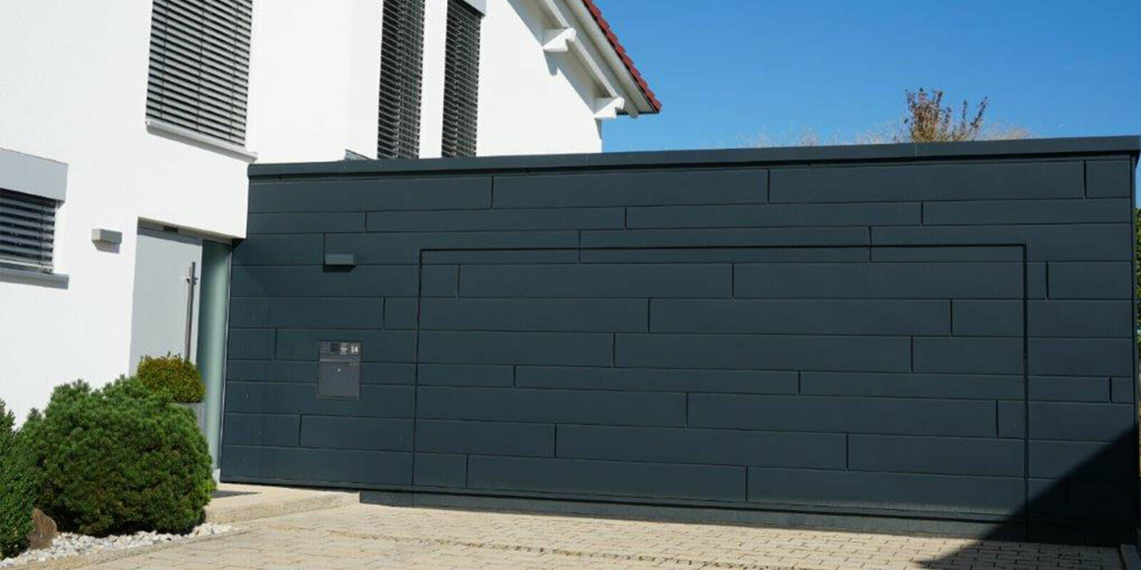 Façade d’un garage après la rénovation à l’aide de Sidings PREFA couleur anthracite. La porte de garage est alignée avec la façade